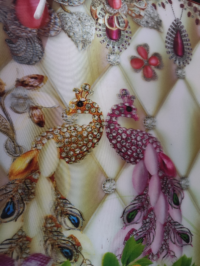 This image depicts a decorative, multi-colored patterned surface, observed in Astanajapura, Indonesia. The background features a quilted or tufted design in light cream or off-white, forming diamond shapes, each intersection marked by a clear, round, faceted jewel-like depiction. Central to the pattern are two stylized peacocks facing each other. The peacock on the left is predominantly gold-toned, its body adorned with numerous clear, simulated gemstones, and its tail feathers include blue "eyes." The peacock on the right is predominantly magenta-pink, its body also covered in clear, simulated gemstones, and its tail feathers feature green "eyes." Above and around the peacocks, various ornamental motifs resembling elaborate brooches or pendants are visible, incorporating large red or maroon teardrop and floral shapes, surrounded by smaller clear, simulated gemstones, alongside silver-toned intricate designs. Green leaf-like elements are present along the bottom edge. No human subjects, specific actions, or direct interactions are depicted. The environment is an abstract decorative print on a flat surface, possibly a textile or wall covering.