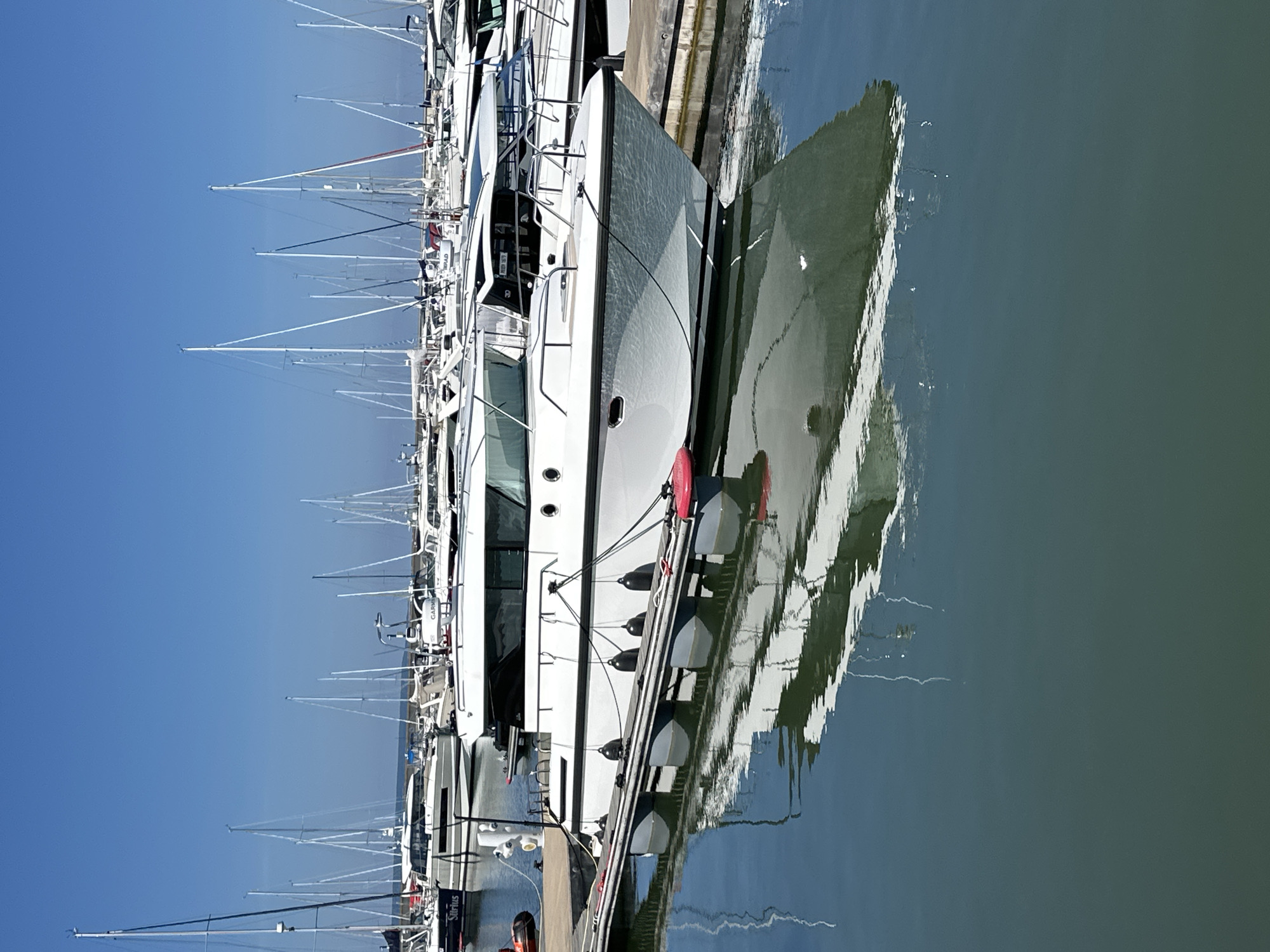 The image depicts a marina scene under a clear blue sky. In the foreground, a white motor yacht is docked at a pier, its reflection visible in the calm water below. The yacht has a sleek design with a cabin, windows, and railings. Several black fenders hang from the side of the pier, and a red bumper is attached to the edge. Behind this primary subject, numerous sailboats with tall masts are moored in the background, creating a picturesque nautical landscape. The overall impression is one of a peaceful and well-maintained marina. The location is identified as the city of Tallinn, Estonia.