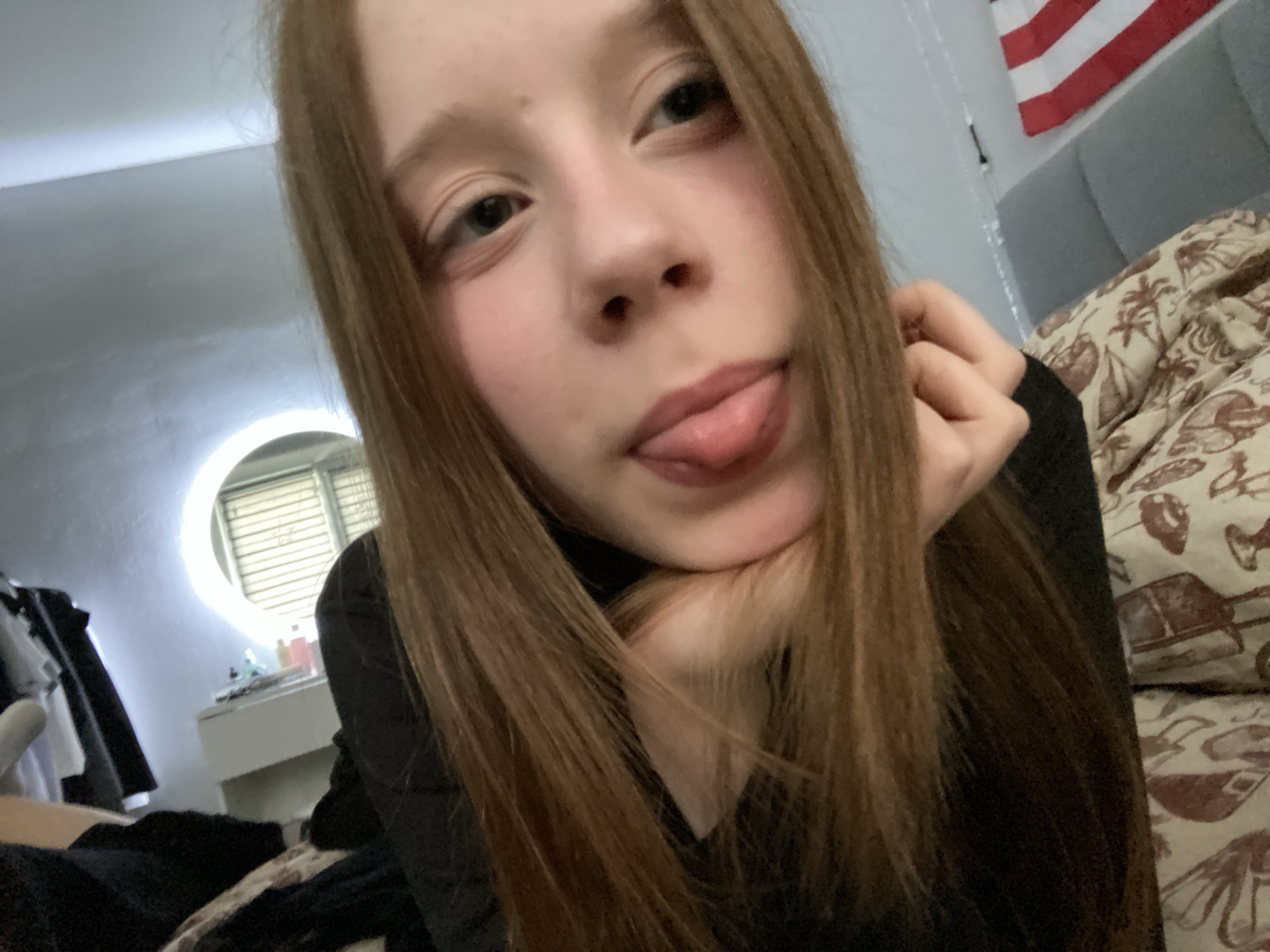 A young person with light skin and long, straight, reddish-brown hair is posing for a close-up selfie. They are wearing a black top and sticking their tongue out. Their face is resting on their hand, and they are looking at the camera. In the background, there is a bed with a patterned comforter, a white dresser with a lighted mirror, and clothing on a rack. An American flag is partially visible on the wall. The location appears to be a bedroom.
