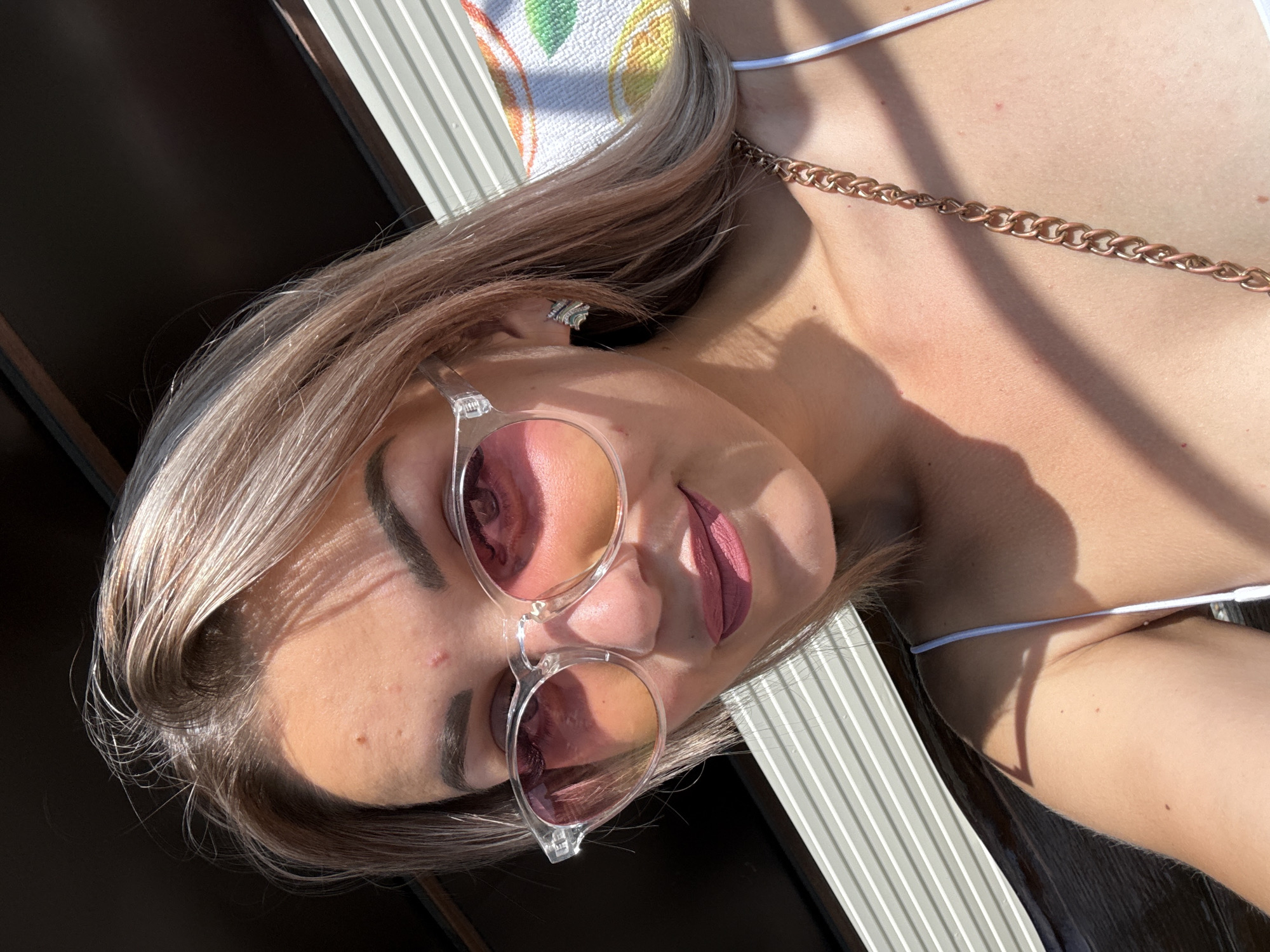 A selfie of a fair-skinned woman with blonde hair and pink-tinted sunglasses is taken outdoors in what appears to be a sunny environment. She is wearing a white top, and a gold chain necklace is visible. The background includes horizontal striped blinds, a dark structure above, and a towel with a fruit pattern. There are subtle imperfections on her forehead. The lighting suggests it is daytime.