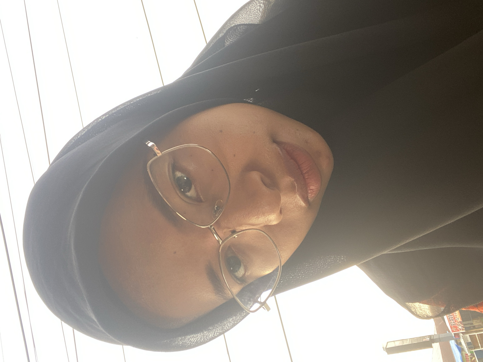 A close-up selfie shows a young woman wearing a black hijab and clear-rimmed glasses. Her face is angled towards the camera, with her right eye looking directly at the viewer. She has a light complexion and her lips are slightly parted. The background consists of a bright, overcast sky with a few thin, horizontal lines that appear to be wires. The overall lighting suggests it is daytime. In the lower left corner, a blurry background shows what appears to be a street scene with buildings and a tall, slender tower. Given the detail, it's likely this scene is within the city of Air Molek, Indonesia.