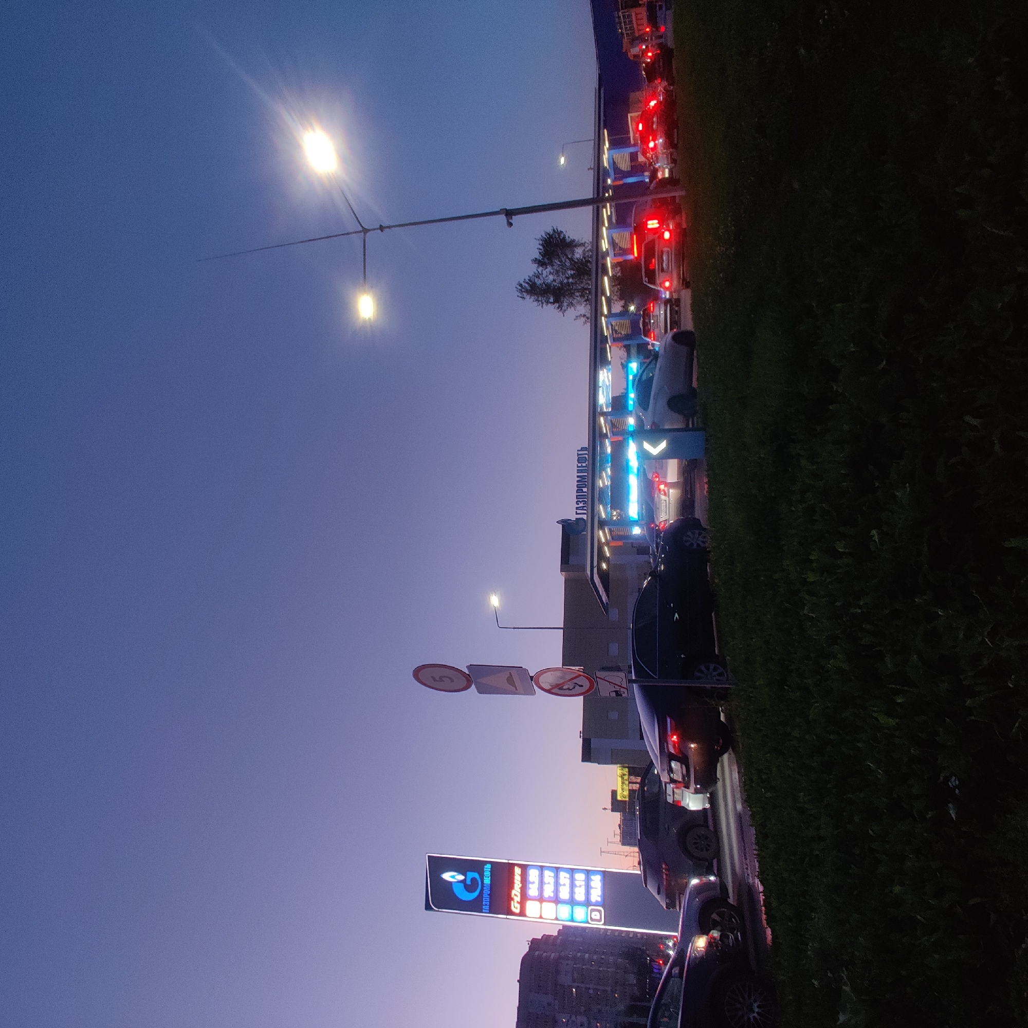 The image captures a twilight scene at a Gazpromneft gas station in Yekaterinburg, Russia. The sky is a gradient of deep blue transitioning to lighter purple hues. A tall, illuminated sign for "GAZPROMNEFT" is prominently displayed on the left, with fuel prices listed on a vertical digital display. Cars are queued at the gas pumps under the covered structure, with their headlights and taillights illuminating the area. A street lamp with two lights is visible in the upper right quadrant, casting a bright glare. Several cars are on the road in the foreground, some parked and others in traffic. Road signs are visible on the right side of the image, including a speed limit sign with a "5" and other regulatory signs. The overall environment suggests a commercial area within the city, with buildings visible in the background. The image is taken from a low angle, focusing on the gas station and the vehicles.