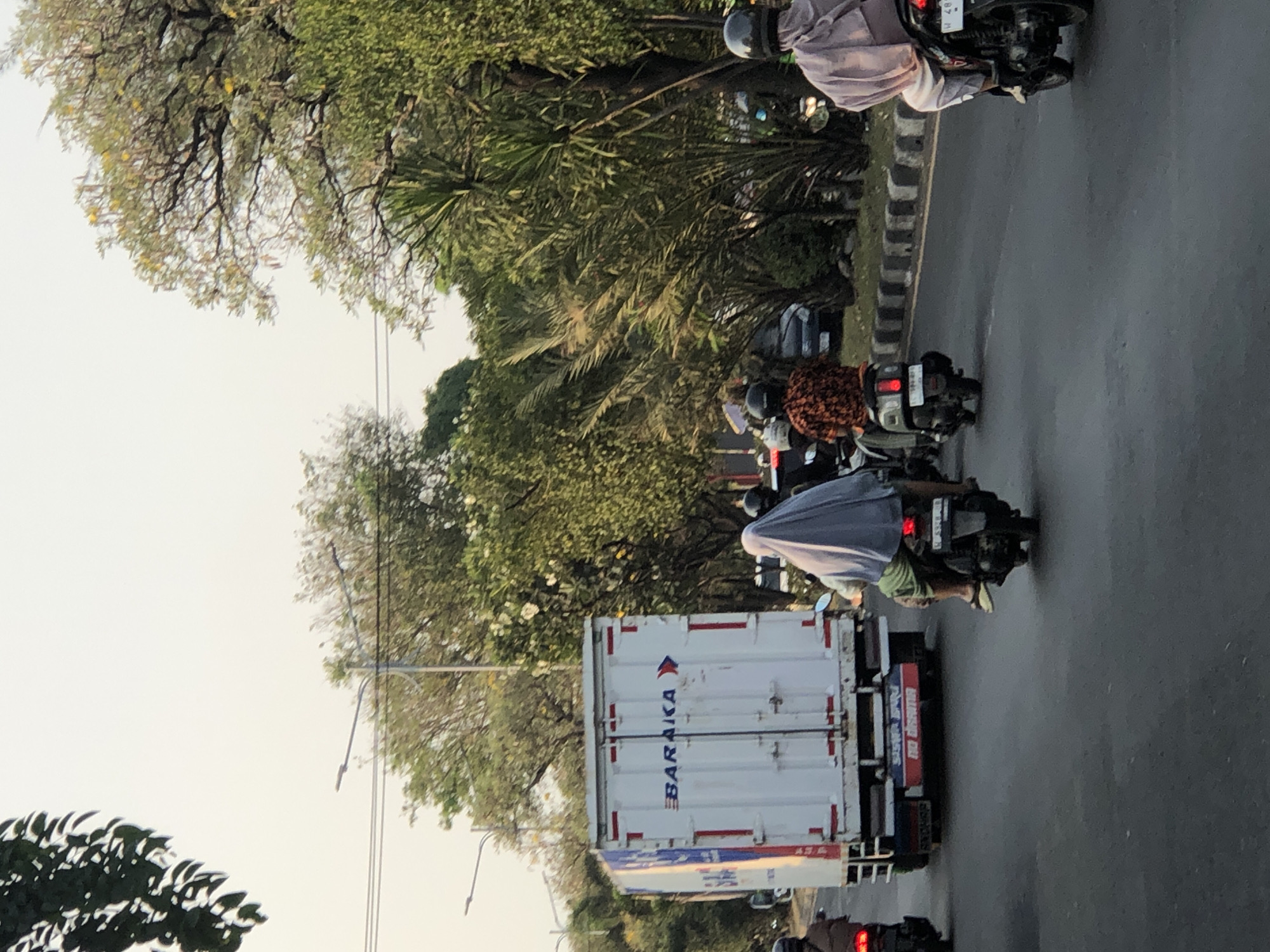 The image shows a street scene in Ngaban, Indonesia, with several vehicles and people. In the foreground, a white truck with "BARAKA" written on its side is driving on a paved road. Behind the truck, two people are riding a motorcycle, the passenger wearing a light purple headscarf and a light-colored garment. To the left of the truck, another motorcycle with a rider wearing a brown jacket is visible. Further down the road, a person on a motorcycle wearing a patterned shirt is seen. In the background, lush green trees line the street, and the sky is overcast. The scene appears to be during daylight hours, possibly late afternoon given the soft lighting. The overall impression is of a typical urban street with traffic.