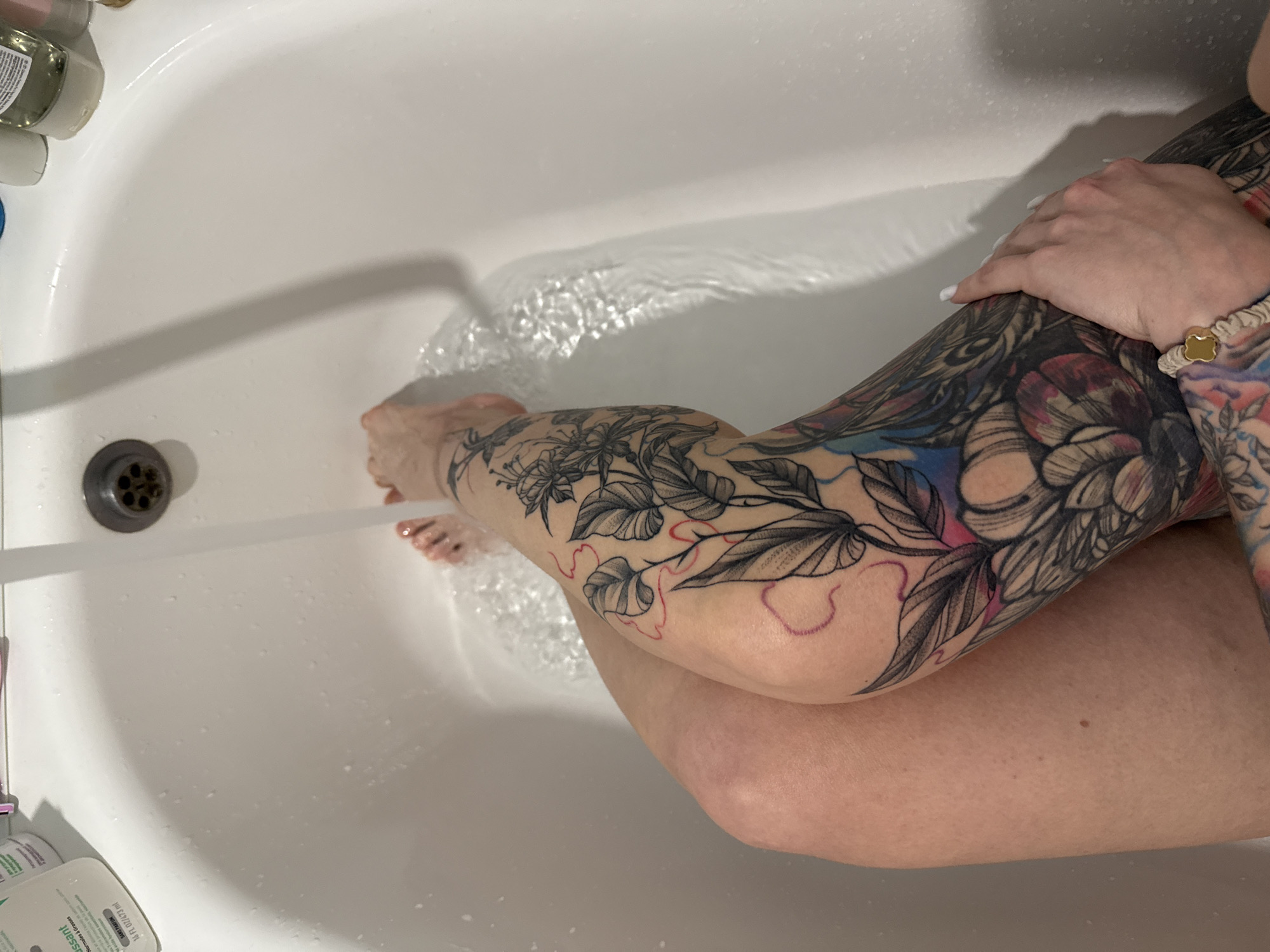 The image shows a person's legs in a bathtub, with water running from the faucet. The person has several tattoos on their legs, including a floral design. The bathtub is white and appears to be in a home bathroom. There are several bottles of toiletries on the edge of the bathtub. This image does not provide any information about the city of Almaty, Kazakhstan.
