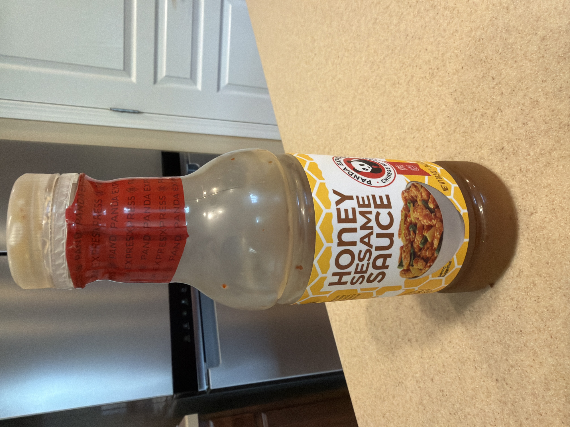 The image shows a bottle of Panda Express Honey Sesame Sauce standing on a speckled countertop. The bottle is mostly clear, with a light brown liquid inside. The label is predominantly white and yellow, featuring the words "HONEY SESAME SAUCE" in large, bold, dark letters. A smaller circular logo with a panda and the words "PANDA EXPRESS CHINESE KITCHEN" is also visible. A picture of a serving suggestion, showing chicken and vegetables in the sauce, is present on the lower part of the label. A red band with repeating text, likely the brand name, wraps around the neck of the bottle. In the background, a stainless steel refrigerator and a white door are partially visible. The scene appears to be indoors, possibly in a kitchen. The provided context states the location is Carpentersville, United States of America.