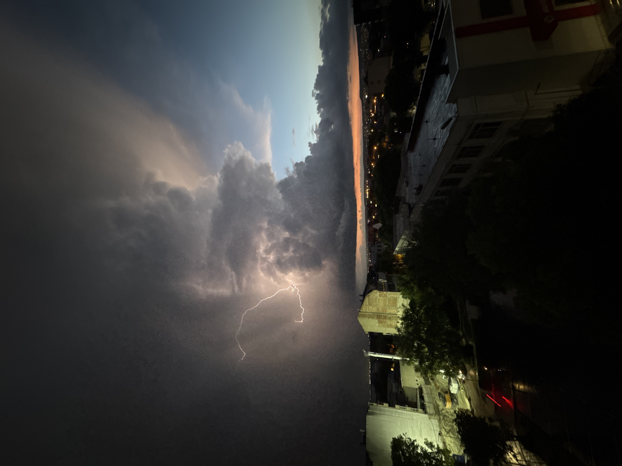 The image depicts a nighttime lightning storm over an urban area, potentially the city of Varna, Bulgaria. A lightning bolt is prominently visible in the center-left of the frame. In the background, there's a city skyline, possibly Varna's, with illuminated buildings and streetlights. The sky is dark with heavy clouds. The foreground shows rooftops and trees, indicating the perspective is likely from a higher vantage point, such as a rooftop or high-rise building.