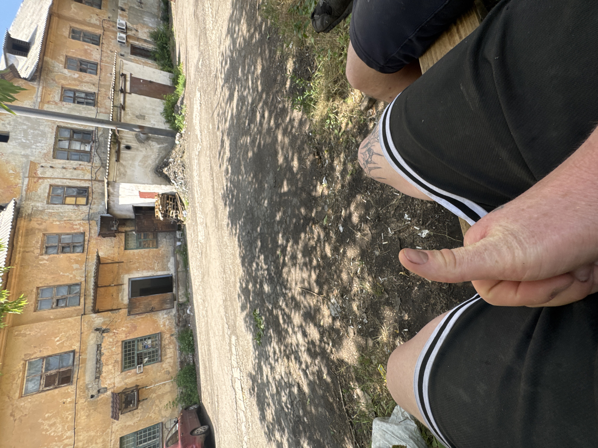 In a courtyard with cracked asphalt, a person's hand gives a thumbs-up gesture. The person is wearing black shorts with white stripes. Behind them, a dilapidated building with peeling yellow paint and broken windows stands under a dappled sunlight. The building appears to be old and possibly abandoned. There is some debris on the ground, including scattered cigarette butts. A red car is parked in front of the building. The overall scene suggests an urban environment in a state of disrepair.