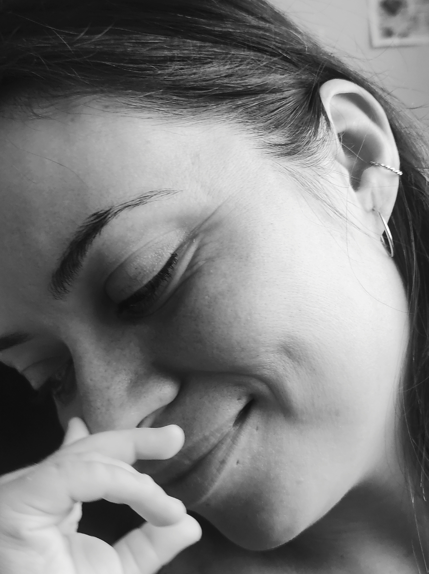 A black and white, close-up portrait captures a person, likely female, in a moment of apparent joy or amusement. The subject's head is tilted slightly downwards and to the left, with eyes gently closed and a smile evident from the crinkling around the outer corner of the right eye. Her right hand is partially raised, covering a portion of her mouth and nose, suggesting shyness, an attempt to stifle laughter, or a candid, heartfelt reaction.
Her dark hair, appearing to be pulled back, frames the right side of her face, with a few wisps visible near her ear. Her right ear is adorned with two delicate silver earrings: a twisted band piercing the upper cartilage and a simple hoop in the lobe. A distinct, well-defined eyebrow is visible above her closed eye, and her eyelashes are long. The lighting appears soft, highlighting the contours of her face and showing subtle texture in her skin, including faint freckles on her nose and cheek.
The background is softly blurred, providing minimal environmental context, though a hint of a framed object is barely discernible in the upper right. The image lacks specific location cues for Chaikovsky, Russia, but the intimate framing and expression convey a personal and emotional moment. No text is visible in the image.