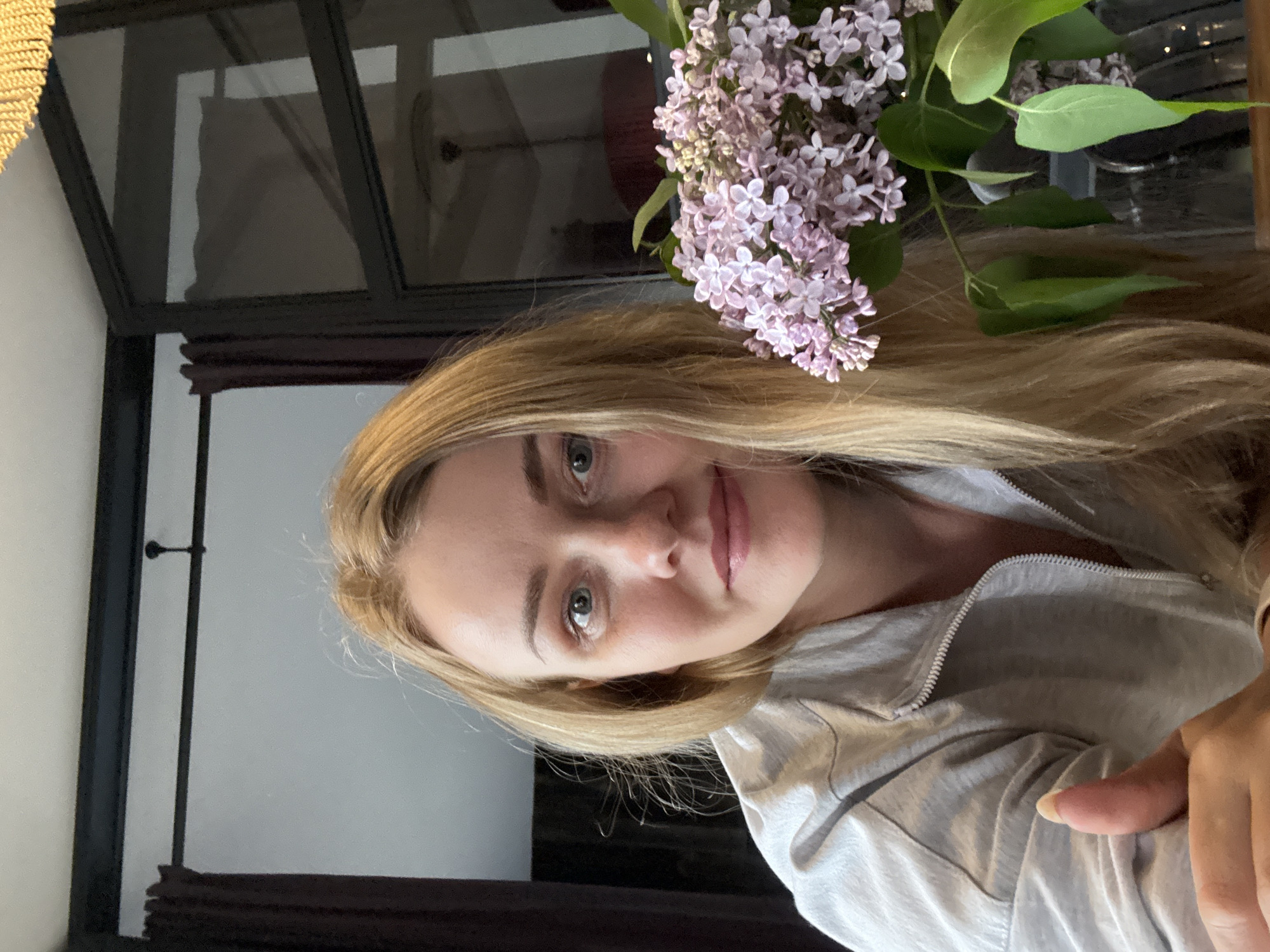 The image shows a fair-skinned woman with blonde hair and light-colored eyes, posing at an angle with a vase of light purple lilacs to her right. She is wearing a light gray zip-up jacket. The background features a dark-framed window or partition, suggesting an interior space. The location cannot be determined as Saint Petersburg, Russia, based solely on the visual information provided.