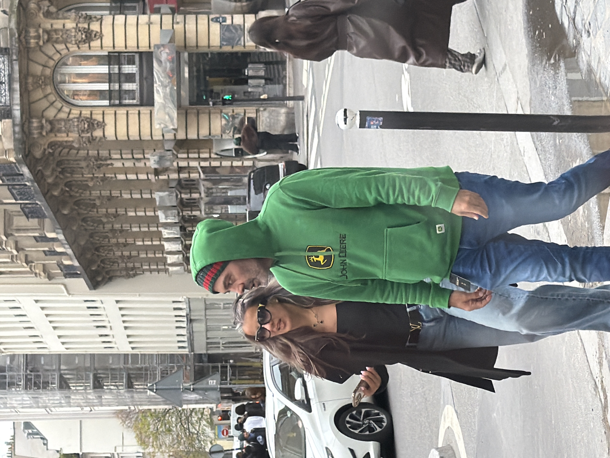The image depicts a street scene in Paris, France. A couple walks along the sidewalk. The man wears a green John Deere hoodie and blue jeans, and a multi-colored beanie. The woman wears a black top, a black coat, and sunglasses. She holds what appears to be a cell phone in her hand. They walk past a white car parked along the curb. In the background are buildings with Parisian architecture. A traffic signal is visible in the distance.