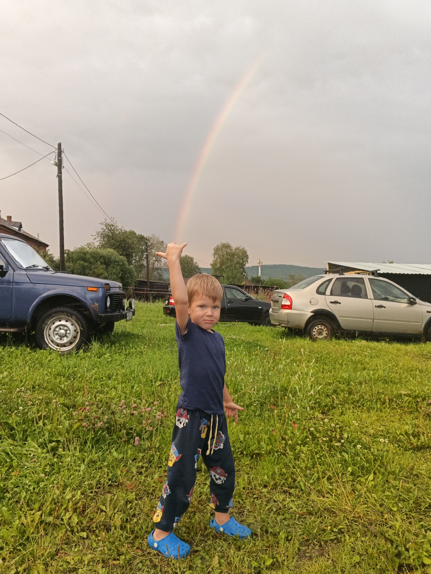 A young boy stands in a grassy field, his right arm raised and bent at the elbow, with his thumb and pinky finger extended in a shaka gesture. He wears a dark blue t-shirt and dark pants adorned with colorful cartoon characters, along with bright blue Croc shoes. Behind him, a light silver sedan is parked, and to the left, a dark blue SUV is visible. A faint rainbow arcs across the overcast sky. The scene appears to be in a rural or semi-rural area, possibly a backyard or open lot, with trees and a distant building in the background. The lighting suggests it may have recently rained or is about to.