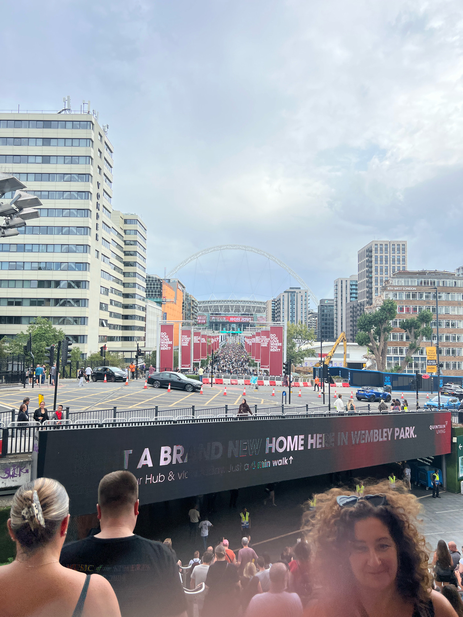 The image captures a busy urban scene in Wembley Park, London, United Kingdom. In the background, the iconic Wembley Stadium arch is visible, along with numerous apartment buildings and city infrastructure. A large crowd of people is gathered, moving towards the stadium or the surrounding area. A prominent digital billboard displays a message advertising "A BRAND NEW HOME HERE IN WEMBLEY PARK."
In the foreground, several individuals are visible from behind, suggesting they are also part of the crowd or observing the scene. The atmosphere appears to be one of an event or gathering, possibly a concert or sporting event given the proximity to Wembley Stadium. Traffic cones and temporary barriers indicate crowd management and traffic control measures in place. The sky is overcast with scattered clouds, suggesting typical London weather.