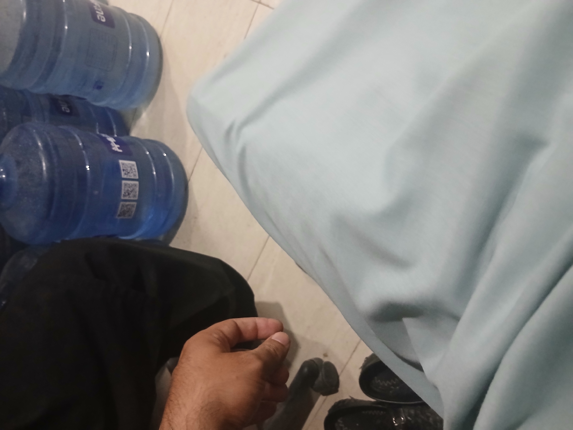 A close-up, indoor shot shows several large, translucent blue water bottles with white labels. The bottles are arranged in a row on a light-colored tiled floor. One bottle in the foreground has QR codes on its label. In the lower left corner, a hand with visible hair on the forearm is resting near a black shoe. To the left of the hand, a black fabric is visible, possibly clothing. In the foreground, a light blue fabric, possibly a blanket or clothing, is draped over a surface. The overall impression is of a casual, domestic setting in Peshawar City Tehsil, Pakistan.