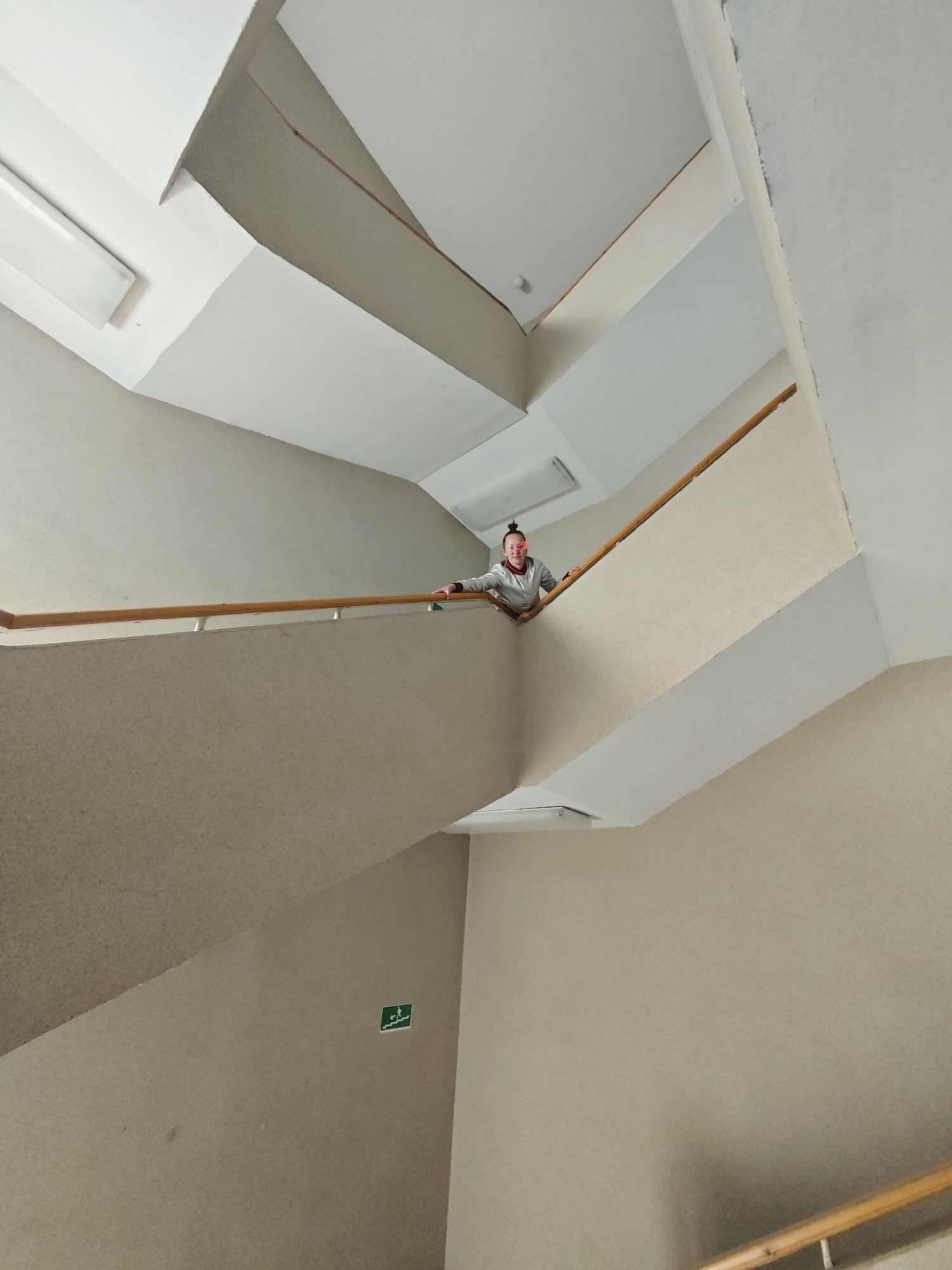 A medium shot captures a person on the landing of a stairwell. The person is fair-skinned with their hair styled up, wearing a light gray zip-up jacket. The person's left arm is visible over the wooden railing. The stairwell walls are a neutral beige color with a green emergency exit sign visible on one wall. The ceilings and some wall sections are white, with fluorescent lights installed. The location is unknown, and the presence of the city of Марковский, Russia is not verified.