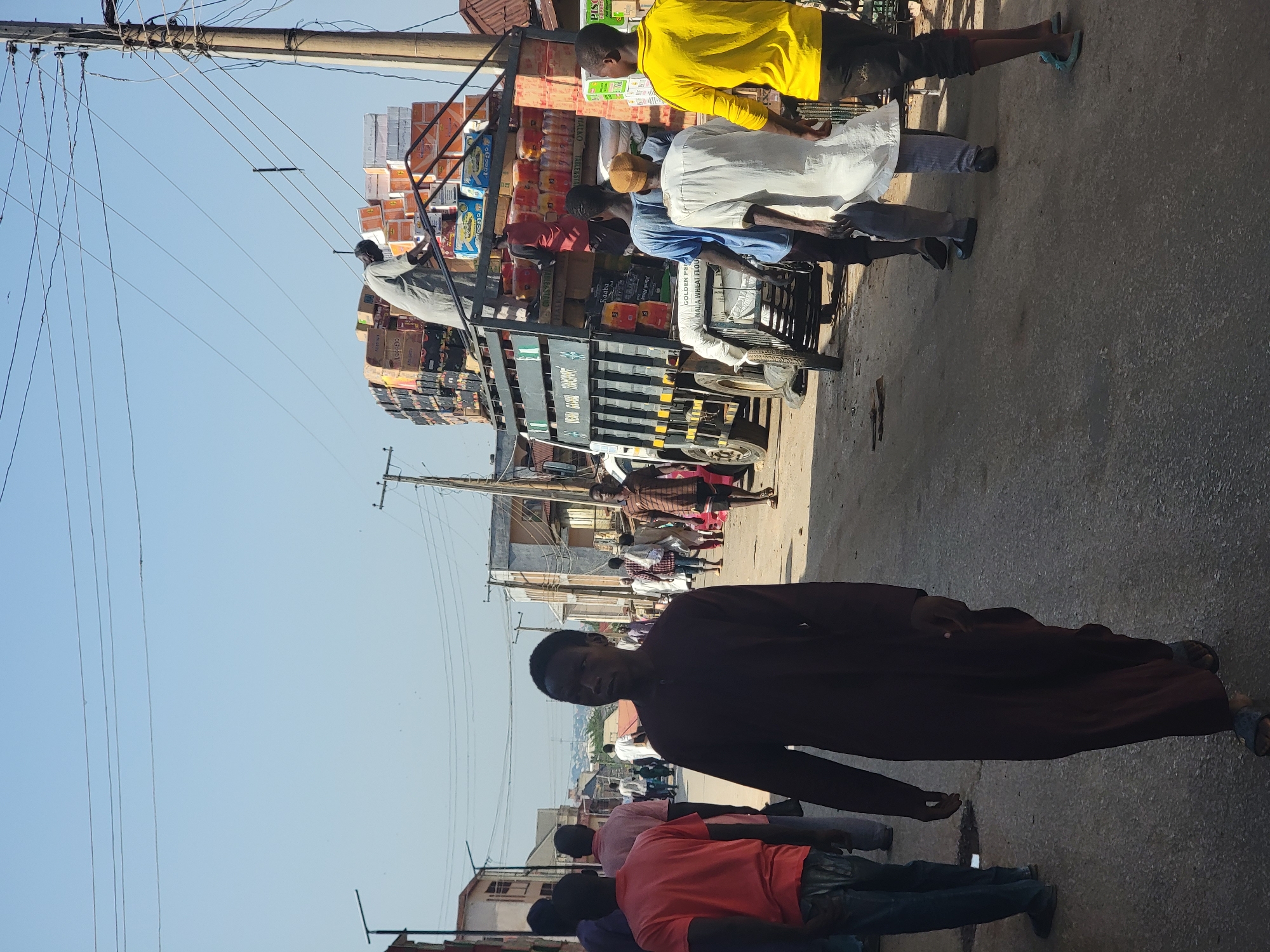 The image depicts a street scene in Maiburiji, Nigeria. A truck laden with stacked goods is parked in the road with people standing around it. Some individuals appear to be loading or unloading the truck. People are also walking along the street. Overhead, power lines crisscross the sky. Buildings are visible in the background.