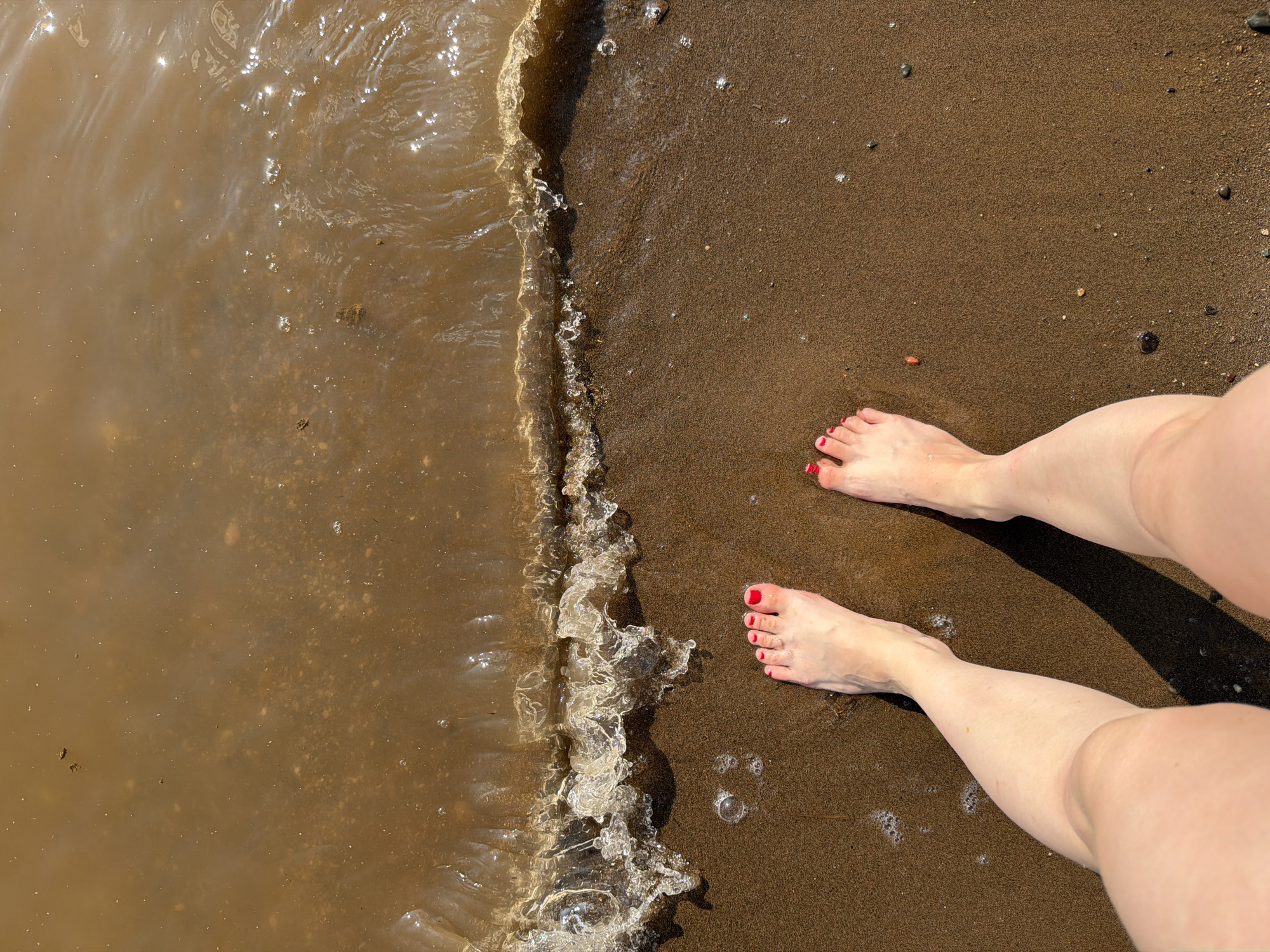 The image shows a pair of bare feet with red nail polish standing on a sandy shore. A wave is breaking at the edge of the water, creating white foam. The water appears murky and brown. The sand is dark brown and wet where the wave has touched it. The scene is likely a natural setting, possibly a riverbank or lake shore, in Krasnyi Bor, Russia. The lighting suggests it is daytime, and the sun is casting a shadow to the right of the feet. There are no other people or objects visible in the frame.