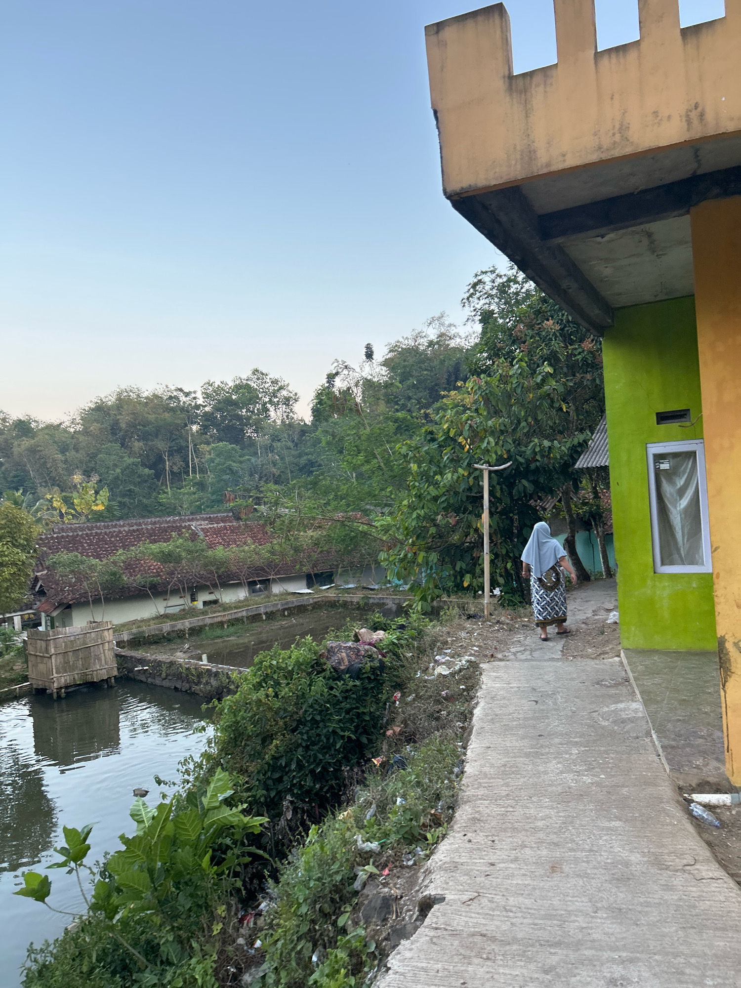 The image shows an outdoor scene with a body of water, buildings, and vegetation. In the foreground, a concrete pathway runs alongside a green and yellow building. A woman wearing a headscarf and patterned skirt walks away from the camera on the pathway. To the left of the pathway is a body of water with some bamboo structures at its edge. Beyond the water, there is a building with a red-tiled roof and a hill covered in trees in the background. The sky is clear and light blue. The scene appears to be in a rural or semi-rural area, possibly a village or a fish farm, given the water and the presence of what might be fish ponds. The overall impression is one of a quiet, everyday scene in a tropical or subtropical environment.