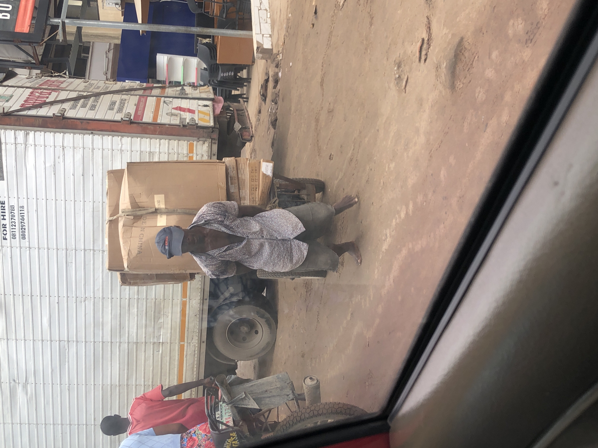 The image depicts a street scene in Lagos, Nigeria. A man wearing a patterned button-down shirt, shorts, and a baseball cap is standing next to a handcart laden with cardboard boxes. The man appears to be in the process of moving the boxes. To the left of the man, another individual in a red t-shirt is visible. Behind them is a large white truck with "FOR HIRE" and phone numbers printed on its side. The ground is unpaved and dusty, indicating a typical street condition in Lagos. In the background, there are buildings and what appears to be an open market or shop area. The perspective suggests the photo was taken from inside a vehicle, possibly a car, looking out through the window.