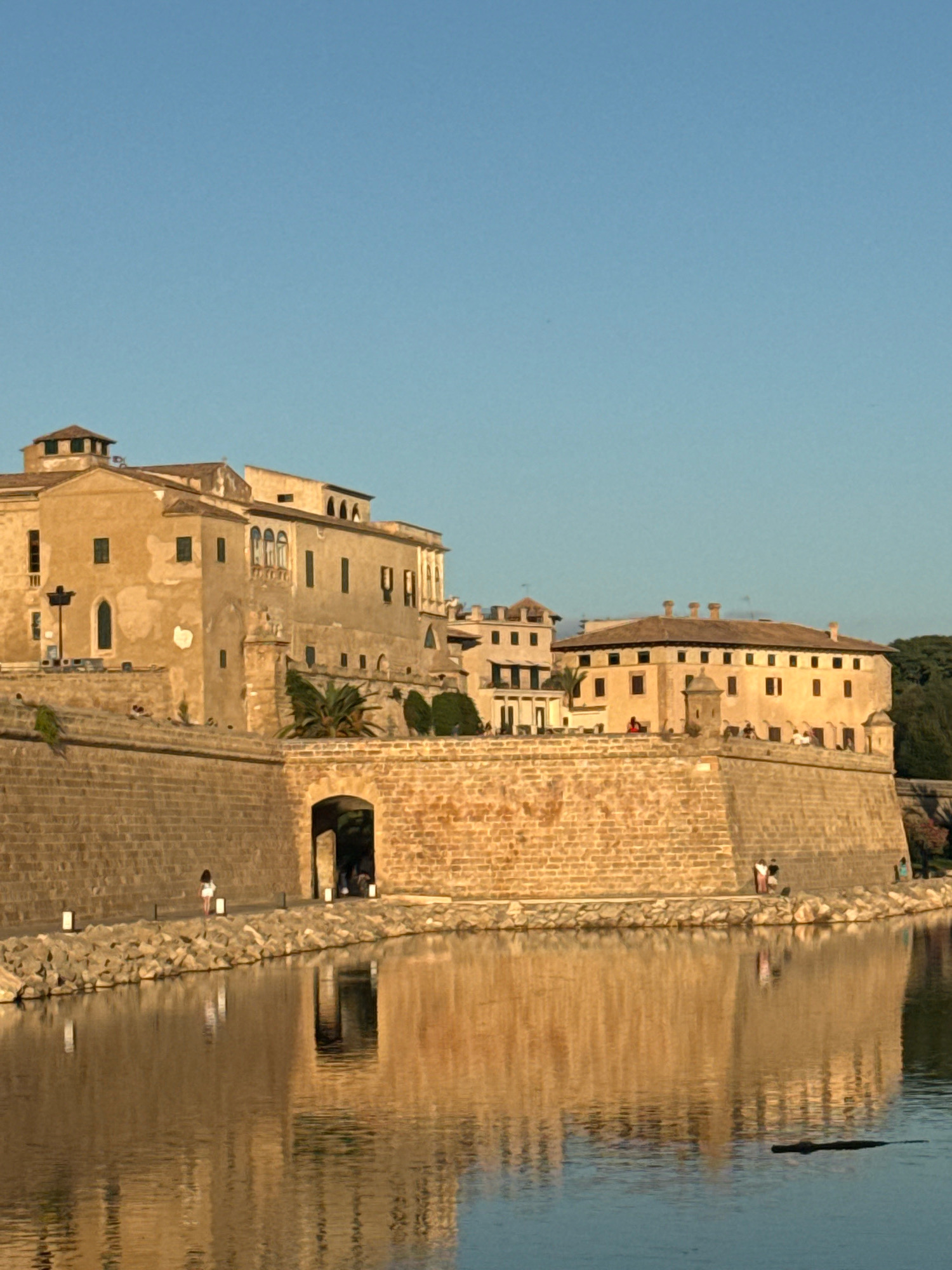 The image shows a waterfront view of the historic Castell de Sant Carles in Palma, Spain, under a clear blue sky. The castell, a large stone fortress, is situated on a raised embankment overlooking a body of water. The stone walls of the castell are weathered and a light sandy color, with arched openings and a prominent gateway visible. Several buildings with terracotta roofs and tan facades are perched atop the castell walls. Green vegetation, including a palm tree, grows on the ramparts. The water in the foreground reflects the castell and the sky, creating a serene and picturesque scene. A few individuals are visible on the path along the castell wall, and one person stands near the gateway. The overall impression is one of historical grandeur and tranquility.