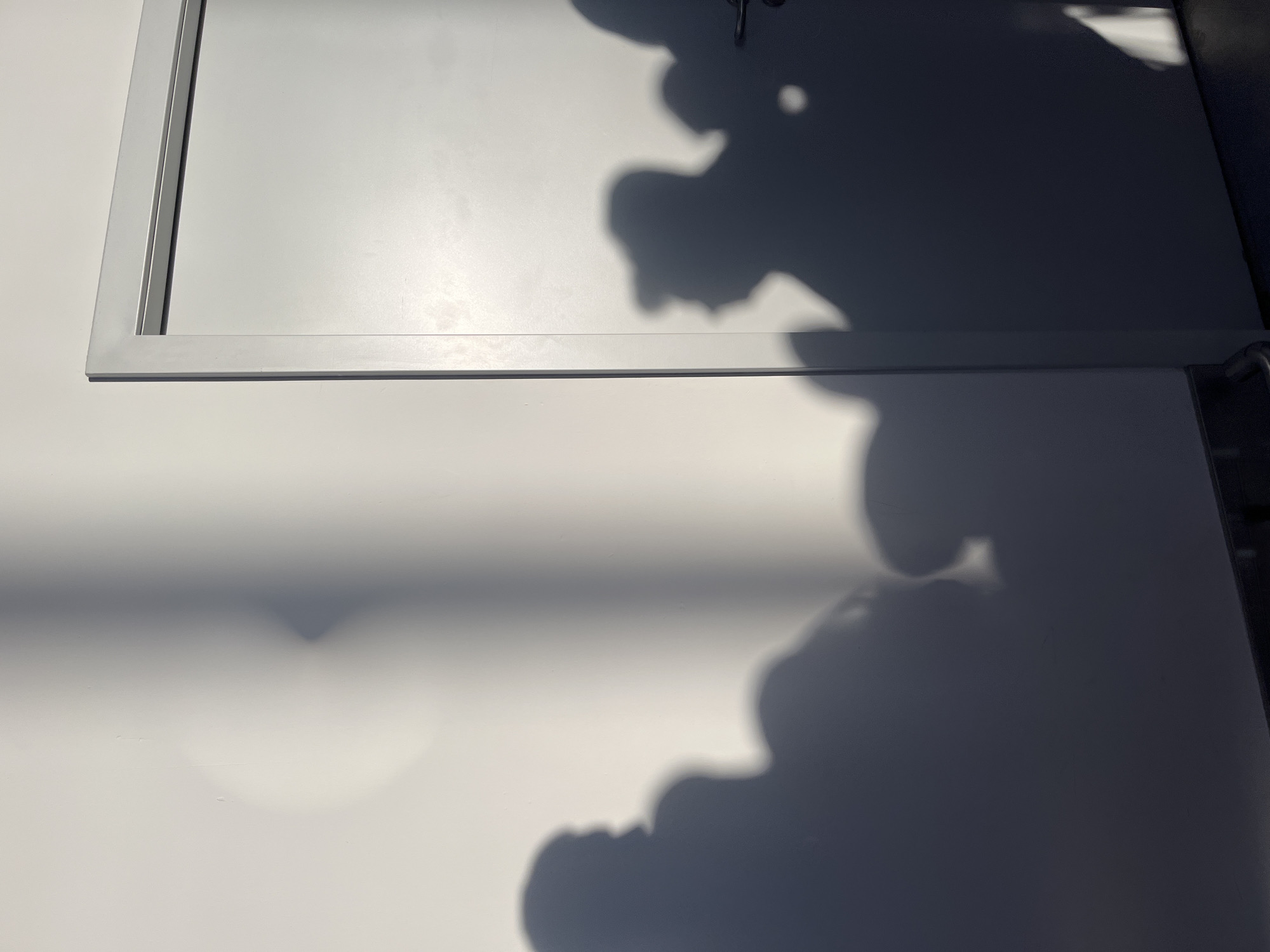 The image shows a wall with shadows cast upon it, to the right of the wall is a white door. The shadows are of several people, some appearing to be wearing hats. The light source is to the left of the frame, casting long shadows. The floor is visible at the bottom of the image, and it appears to be dark and possibly tiled. The wall is painted white or a light color, and the door is also white with a silver handle. The scene appears to be indoors or in a sheltered outdoor area with a smooth wall surface. The location in the city of Aksu, Turkey is not discernible from the image.