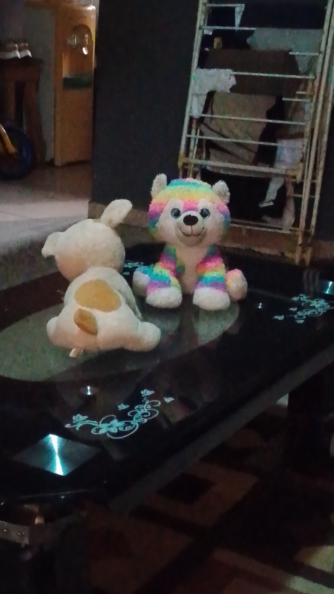 The image shows two stuffed animals on a black glass table. One is a white bunny with brown spots on its back, facing away from the viewer. The other is a rainbow-striped bear, facing forward. A metal drying rack with clothes on it stands against a wall in the background. A water dispenser and what appears to be the front tire of a child's tricycle are partially visible in the left background. The floor appears to be tiled. It is difficult to determine the specific location within Asubi, Nigeria based solely on this image.