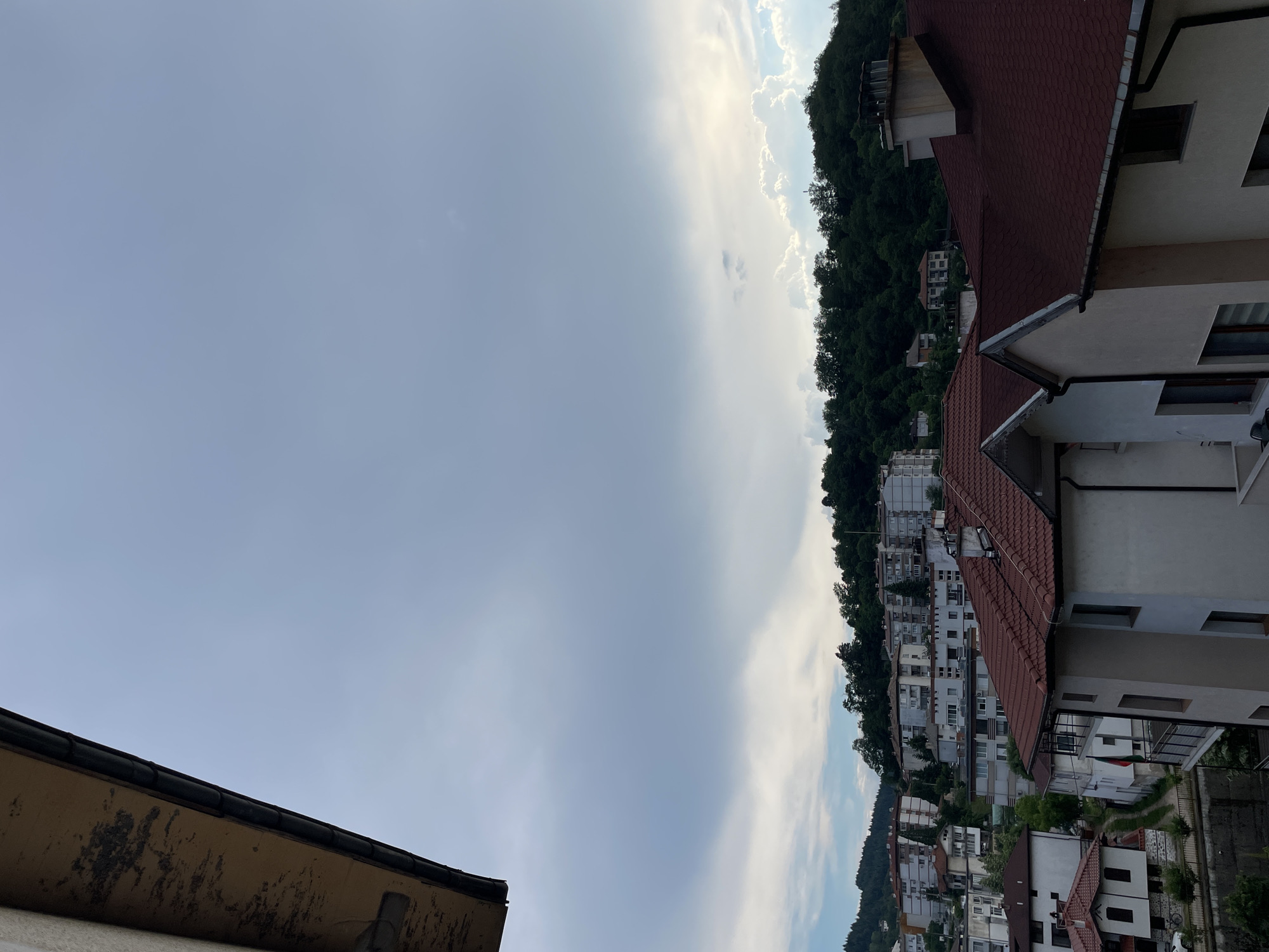 This media file captures an overcast sky above a residential area nestled on a hillside. The scene is dominated by a gradient of sky, transitioning from a pale, hazy blue on the left to brighter, cloud-filled expanses on the right, with the sun's glow faintly visible through the clouds. The hillside on the right is densely covered with dark green trees, creating a stark contrast with the lighter sky.
Below the trees and along the hillside, numerous buildings are visible. Many of these are houses and apartment buildings with red-tiled roofs, and pale-colored facades. Some of the buildings appear older, while others seem more modern. The architecture suggests a European setting. The image is oriented sideways, with the sky occupying the left portion and the cityscape and hillside on the right.
The time of day appears to be late afternoon or early evening, given the soft, diffused light. The weather seems to be cloudy, possibly with a chance of rain or just post-rain. The location is Zlatograd, Bulgaria, as indicated by the context provided. No people are clearly visible, and there are no discernible signs of specific activities or events. The overall mood is calm and somewhat subdued due to the overcast conditions. There is no visible text in the image.