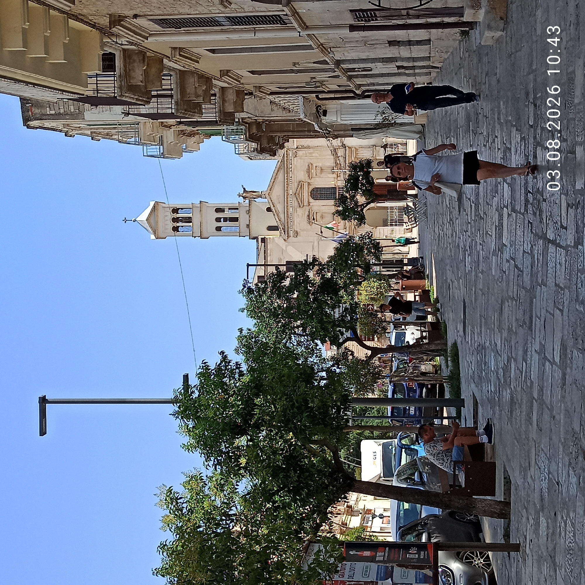 The image depicts a street scene in Ruvo di Puglia, Italy, on August 3, 2026, at 10:43 AM. The perspective is from street level, looking down a cobblestone street lined with buildings and trees.
In the foreground, a man wearing a patterned short-sleeved shirt, blue shorts, and white socks with "FILA" branding sits on a park bench, looking at his phone. He is wearing dark shoes. A tree is positioned behind him. To his left, a silver car is parked.
Further down the street, several people are walking. A woman in a white t-shirt and black skirt walks towards the viewer, carrying a white tote bag. Behind her, a man in a dark polo shirt and jeans walks away from the viewer, holding a book and a drink. Other pedestrians are visible in the mid-ground and background.
On the left side of the street, there are several parked cars, including a white truck with "GLS" branding. A billboard with Italian text is also visible on the left.
Dominating the background is a light-colored church with a prominent bell tower topped with a cross. Statues are visible on the church facade. To the right of the church, a multi-story building with balconies and arched windows is visible.
The sky is clear and blue. The street is paved with large, irregular cobblestones. The overall scene suggests a sunny day in an urban environment with a mix of residential and religious architecture.