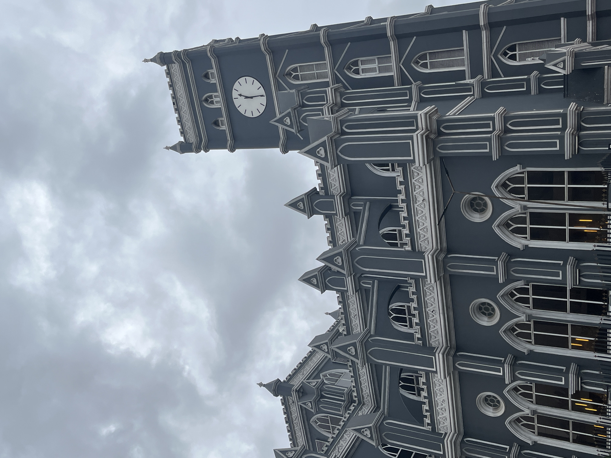 The image depicts a tall, gray cathedral-like structure with white trim, possibly made of stone or concrete. It features a prominent clock tower with a clock face showing approximately 9:15. The architecture is Gothic-inspired, with pointed arches and intricate detailing. The sky is overcast with gray clouds. The angle suggests the photo was taken from below, looking upwards. The location is not specified.
