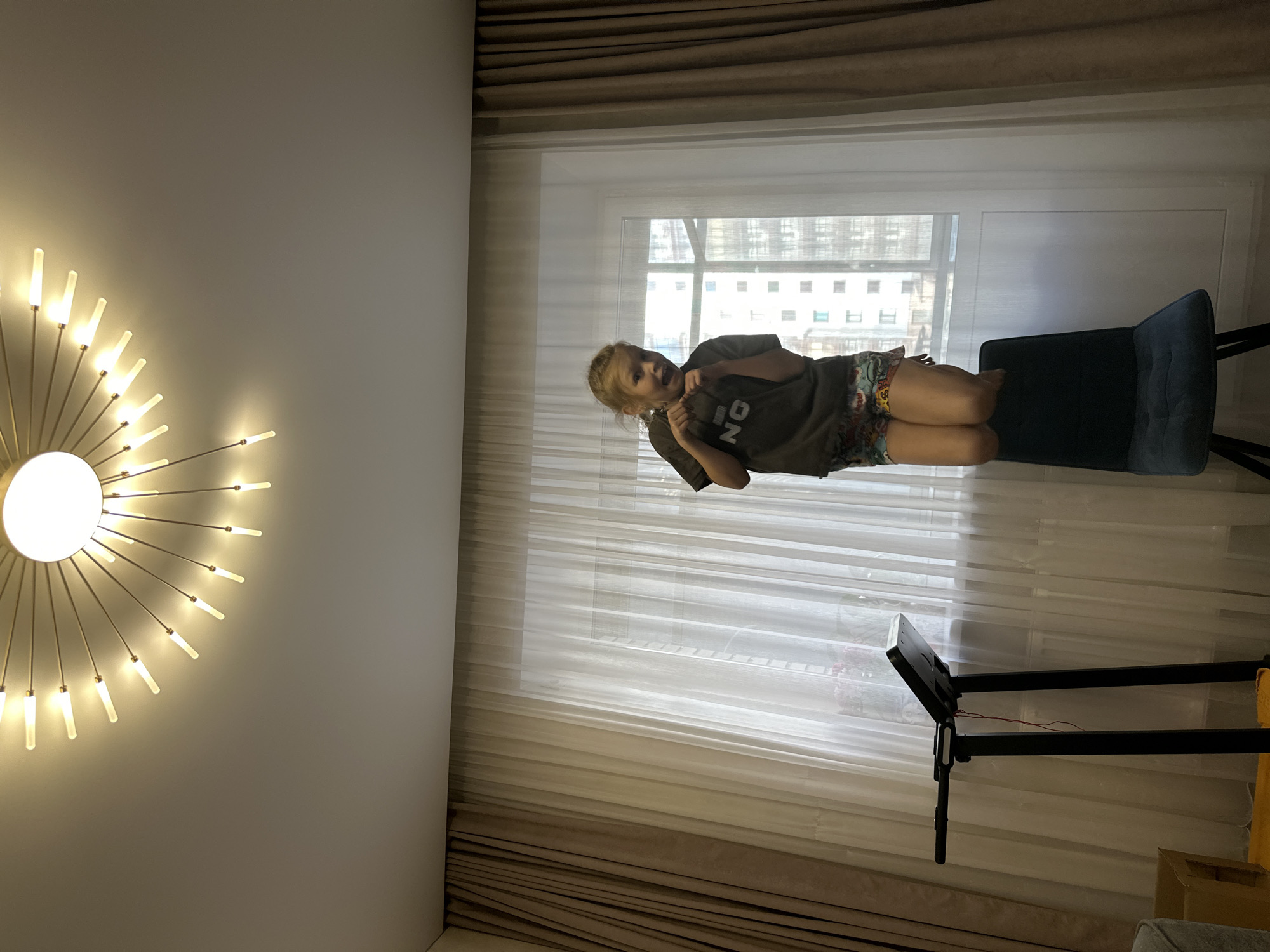 A young girl with blonde hair is standing on a chair in front of a window. She is wearing a dark t-shirt with "NO" printed on it and colorful shorts. Her hands are raised in a playful stance, and her mouth is open in an excited expression. The girl is located in a room with sheer white curtains covering the window, and darker brown curtains on either side. A treadmill is visible to the left of the girl. A modern, starburst-shaped chandelier hangs from the ceiling. The background shows apartment buildings through the window. The scene is set in an interior residential space.