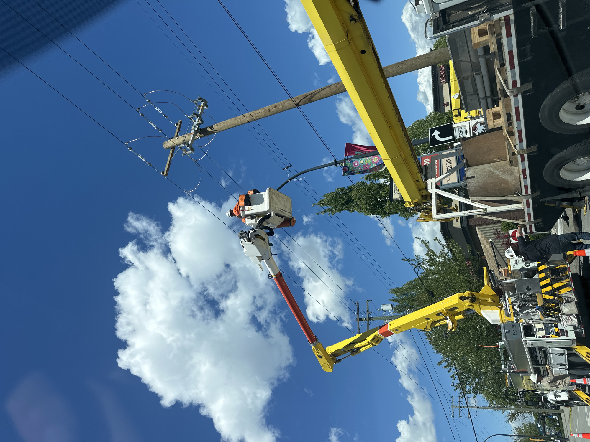 An aerial view shows a bright blue sky with scattered white clouds. A utility truck with a large yellow crane arm is parked on a street. A worker in a white hard hat and orange vest is in the bucket of the crane, working on overhead power lines. Another worker in a white hard hat is standing on the ground next to the truck. The truck is carrying equipment and supplies. In the background, there are trees, buildings, and more utility poles. A sign on a building reads "NEWTON". The location is the city of Surrey, Canada.