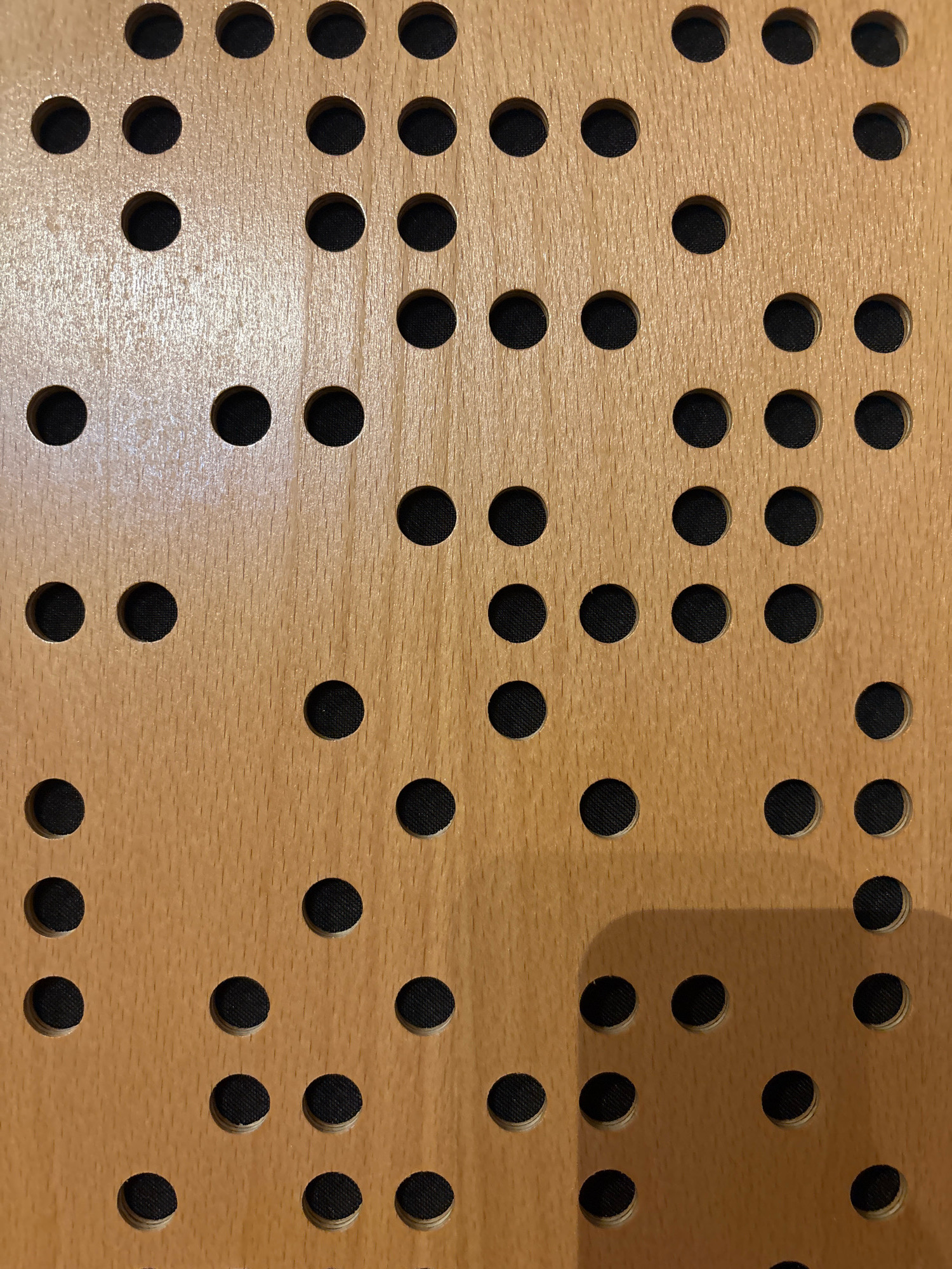 The image displays a close-up, overhead view of a wooden surface with a pattern of evenly spaced, circular holes. The wood has a light brown, grainy texture, and the holes are filled with a dark material, possibly fabric or felt. The lighting creates subtle shadows around the edges of the holes, giving them a slight depth. The scene is devoid of any people, actions, or interactions. The exact location in Chalandri, Greece, is not discernible from the image itself; it appears to be a detail of a manufactured object such as acoustic paneling or a pegboard.