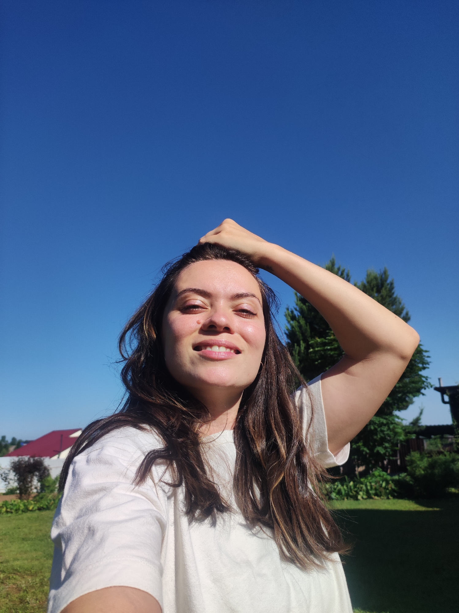 In the city of Fokino, Russia, a young woman with long, dark, wavy hair is pictured outdoors on a sunny day. She is wearing a white t-shirt and has her right arm raised, with her hand resting on her head. The woman is looking towards the camera with a smile. The background shows a clear blue sky, a green lawn, some trees, and a house with a red roof. The image is a selfie taken at a slight upward angle.