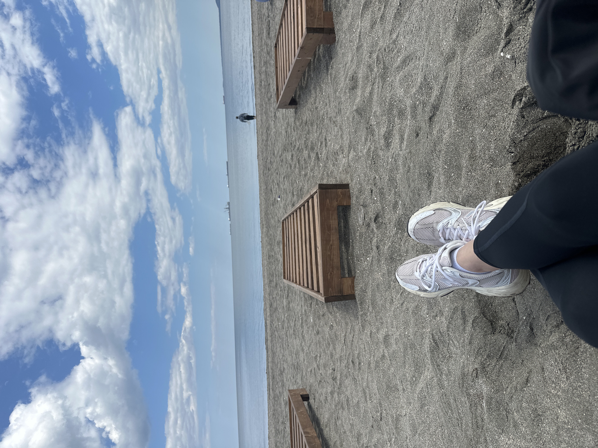 The image, oriented correctly, depicts a serene beach scene from a first-person perspective, looking out towards the sea. In the immediate foreground, a person's legs and feet are visible, clad in dark pants or leggings and white athletic sneakers. The person appears to be sitting or resting on the dark, fine-grained sand.
The beach itself is covered in dark gray sand, possibly volcanic in origin, with scattered small shells or debris. Two wooden beach loungers or low platforms, made of light brown slats, are positioned on the sand, extending towards the water.
Beyond the beach, a vast expanse of calm, light blue-gray sea stretches to the horizon. In the distance, a solitary figure is visible standing in the water near the shore. Faint outlines of what appear to be ships or boats can be seen further out on the horizon.
The sky above is bright and partly cloudy, with significant patches of clear blue visible between fluffy white clouds. The overall impression is that of a clear, bright day, likely late morning or early afternoon. The weather appears pleasant and calm. No specific location cues are visible, nor any discernable text within the image. The scene conveys a sense of peaceful contemplation or relaxation at the seaside.