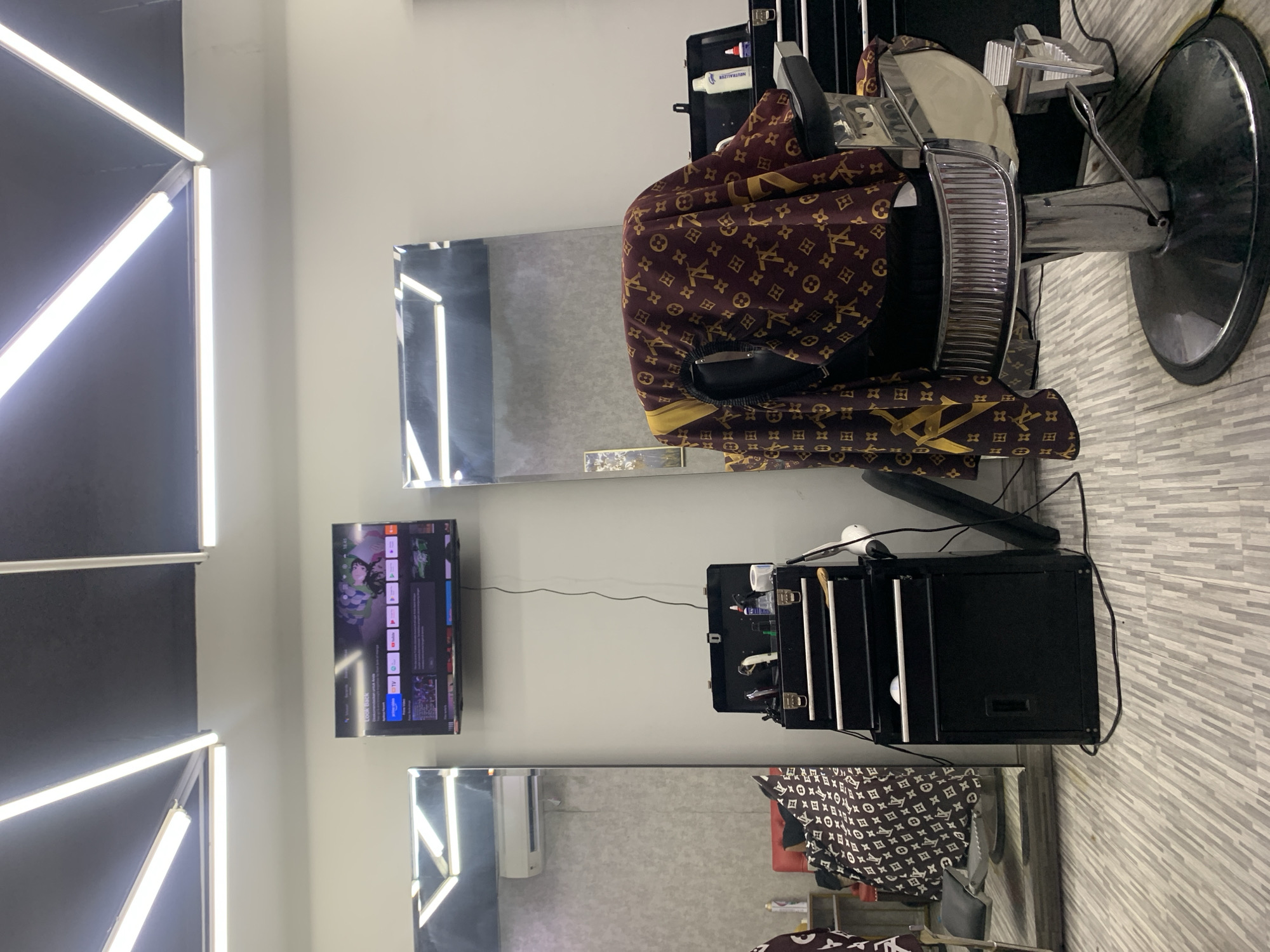 The image displays the interior of a modern barbershop or salon, characterized by a clean and contemporary design. The setting features light grey/white walls, with a distinctive black accent wall on the left adorned with diagonal white LED strip lighting. The floor is tiled with light grey rectangular planks arranged in a horizontal, staggered pattern.
The main subjects are a beige and chrome barber chair positioned on the right side of the frame, and a dark reddish-brown barber cape draped over it. The cape is patterned with repeating "LV" logos and four-leaf clover/flower motifs. A second, similar cape is visible in the reflection of the lower mirror.
Two large, rectangular mirrors are mounted on the wall. Between them, a flat-screen television is mounted, displaying a streaming service interface where "YouTube" and the time "14:43" are visible. To the right of the lower mirror, a black, multi-tiered rolling trolley or portable case is open, containing various barber tools such as hair clippers, combs, and a white tube labeled "NEUTRALIZER." A white hairdryer is also present. Electrical cables run from the TV and the trolley along the wall.
No people are visible, indicating that no specific activity or event is actively taking place, though the setup suggests readiness for haircutting or styling. The time of day is indicated as afternoon by the "14:43" on the TV. The location cues, such as the barber chair and tools, strongly suggest a professional grooming establishment, consistent with a barbershop in Kuta, Indonesia.