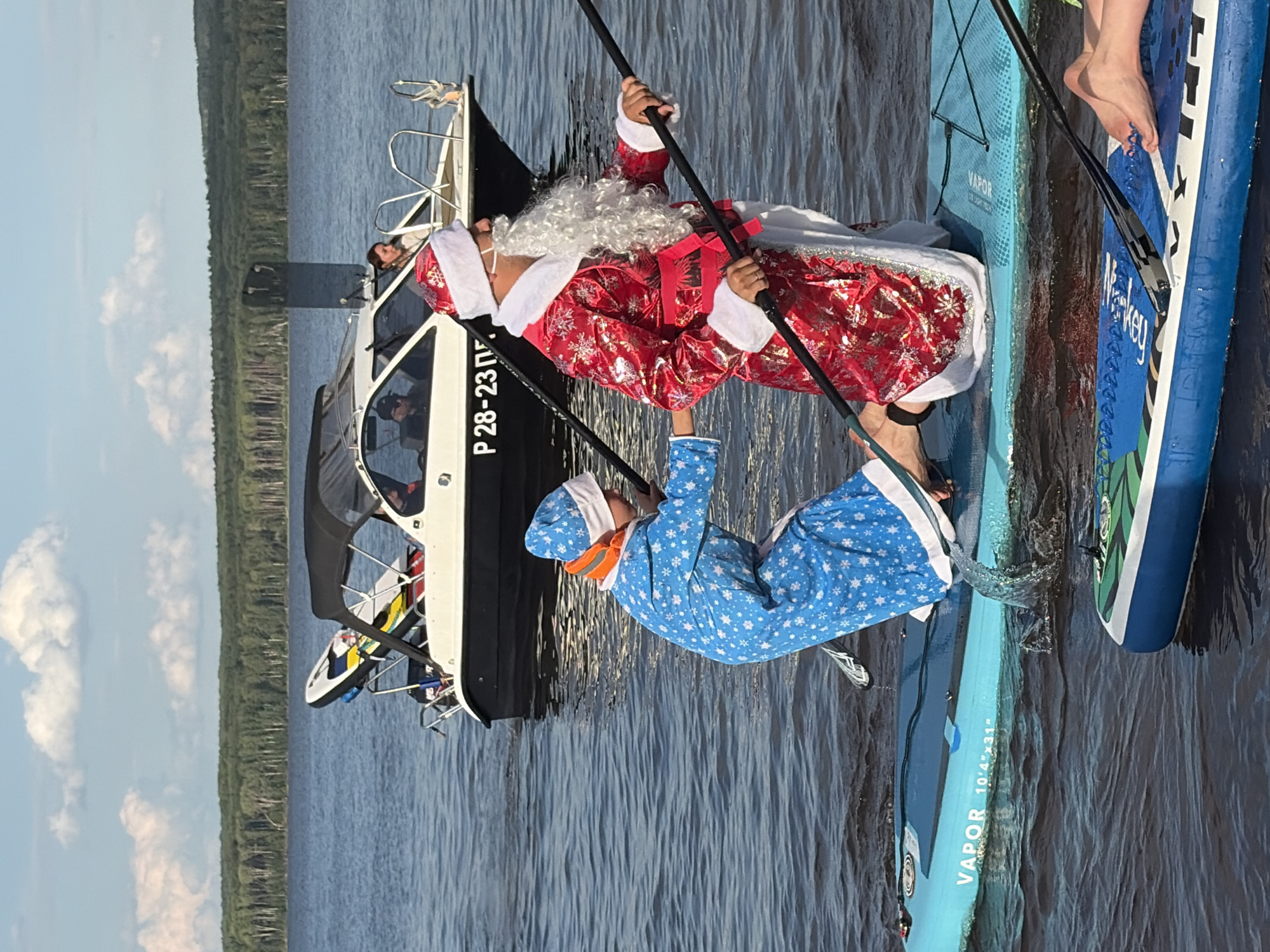 Two people are paddleboarding on a body of water. One person is dressed as Santa Claus in a red suit with white trim and a white beard. The other person is wearing blue pajamas with white snowflakes and an orange neck gaiter. They are both holding paddles and appear to be engaged in a festive activity. The scene takes place on a sunny day, with a boat in the background. Visible text on one of the paddleboards reads "VAPOR 10'4"x31"". The boat has "P 28-23 П" visible on its side. The water is calm, and there are trees and a cloudy sky visible in the distance. The overall mood seems cheerful and fun.