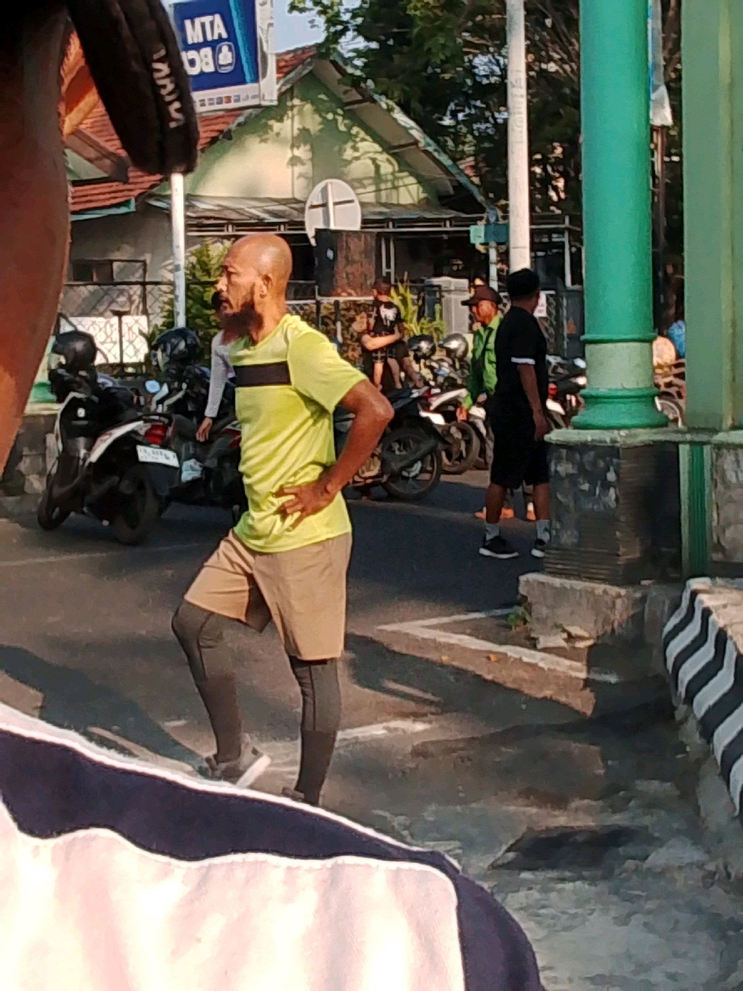 The image depicts a street scene in Probolinggo, Indonesia. In the foreground, a bald man with a goatee is wearing a neon yellow t-shirt with a black stripe across the chest, beige shorts, and dark compression leggings. He is standing with his hands on his hips, looking to his left. Behind him, several motorcycles are parked on the side of the road. Further in the background, other individuals are visible, some appearing to be standing near the motorcycles. The environment includes a building with a green roof, a green pole, and a street with a raised concrete border. A sign with blue and white lettering, possibly advertising a public service or transportation, is also visible. The lighting suggests it is daytime.