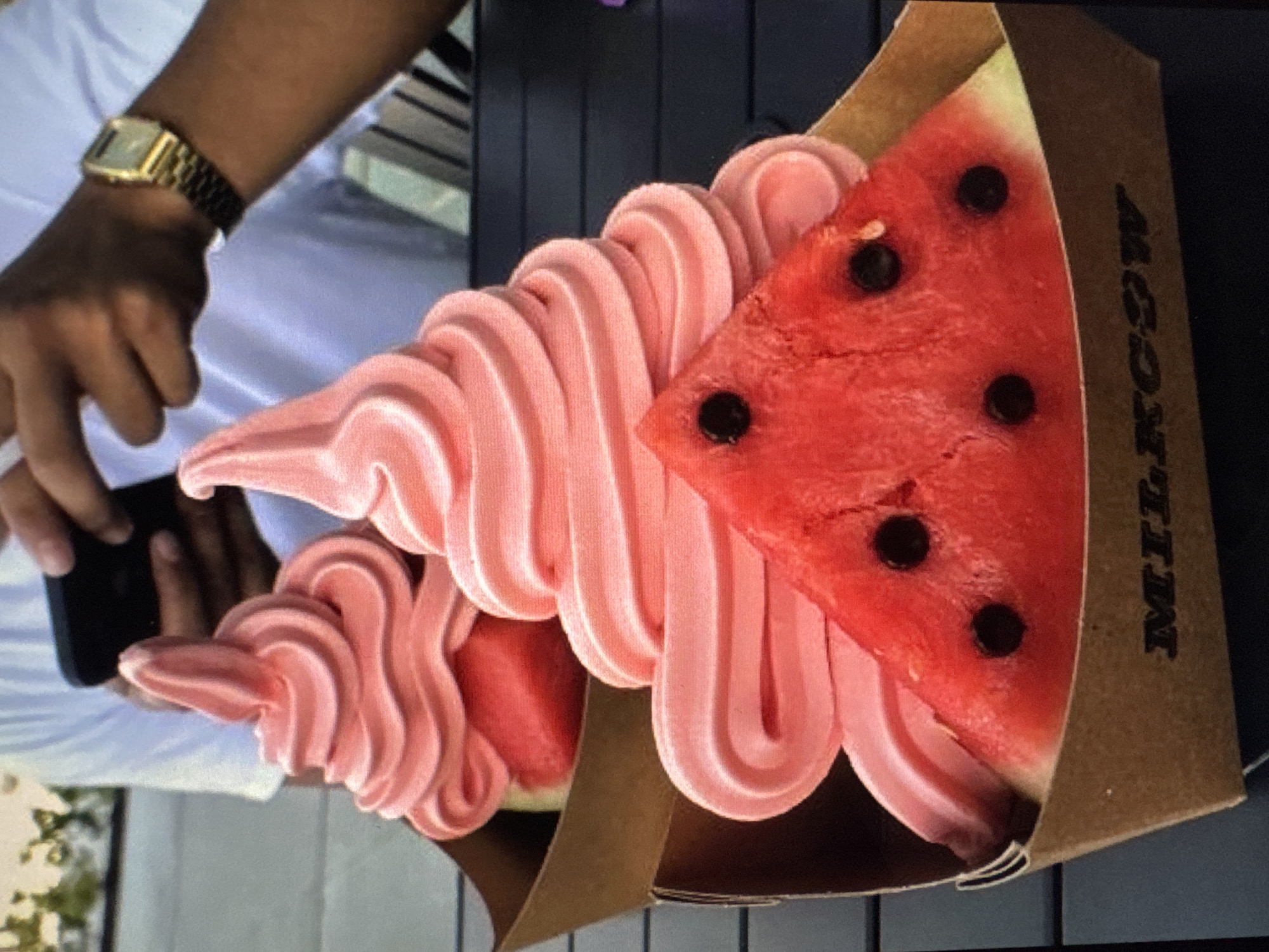 Two pink soft-serve ice creams are presented in brown cardboard containers on a dark surface. Each ice cream is topped with a slice of watermelon, which is triangular in shape and has black seeds. The ice cream in the foreground is larger and more prominently displayed, with its soft-serve swirls clearly visible. The background shows a person in a white shirt holding a smartphone. The scene is set in an outdoor environment, with some greenery and a hint of a building structure in the background. The "MILKCOW" logo is visible on the front of the cardboard container. The specific location within Tallinn, Estonia, cannot be determined from the provided image.