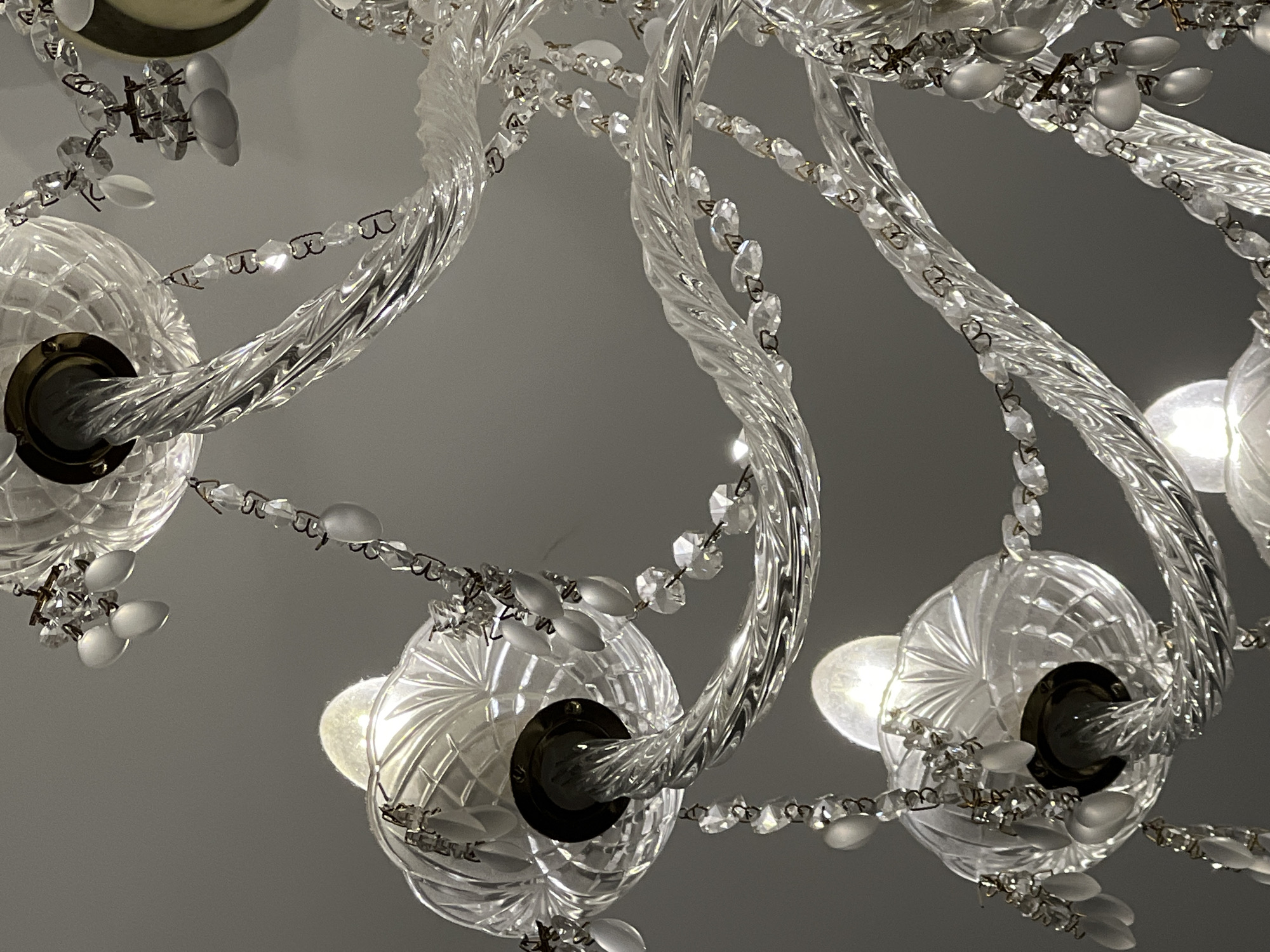 This is a close-up, low-angle shot of a vintage crystal chandelier hanging indoors. The chandelier features twisted glass arms adorned with strings of clear faceted crystals and frosted teardrop-shaped beads. Several light bulbs are visible, glowing softly. The background is a plain, neutral gray, suggesting an indoor ceiling or wall. The image is well-lit, highlighting the intricate details and reflective qualities of the chandelier's glass and crystal components. There are no people, actions, or interactions depicted. The location in Veliky Novgorod, Russia is not discernible from the image itself.