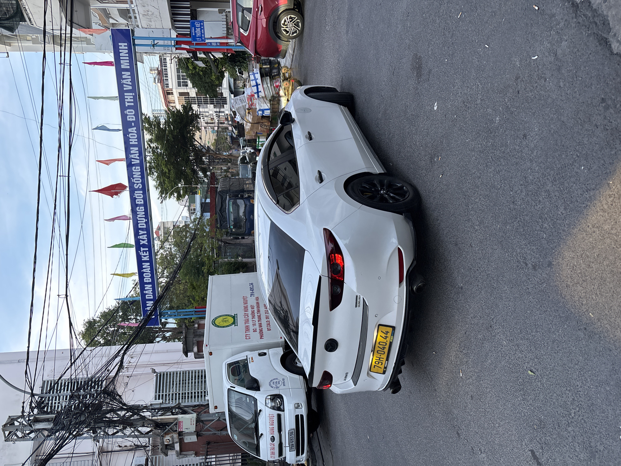 The image depicts a street scene in Nha Trang, Vietnam. In the foreground, a white Mazda sedan is parked, facing away from the viewer, with its rear end prominently displayed. The car has a yellow license plate with the registration number "79H-040.44" and black rims. To the left of the sedan, a white truck with "CTY TINH CAU HUNG NGUYET" printed on its side is parked. The truck has a company logo featuring a green circle with text inside. In the background, a blue banner with Vietnamese text and colorful flags strung across it is visible, suggesting a public event or decoration. The street is paved and appears to be in a commercial or residential area, with buildings and trees lining the sides. There are also other vehicles and objects on the street, including what appears to be a cart laden with goods. The sky is overcast with some sunlight breaking through. The overall impression is a busy urban environment.