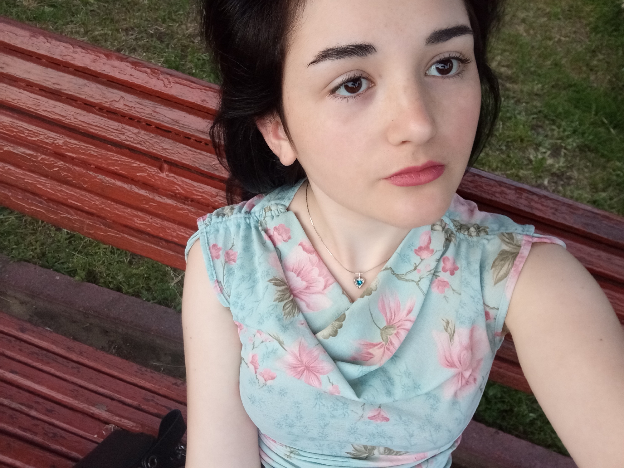 A woman is lying on a red-brown wooden bench. She has dark hair and fair skin. She is wearing a light blue floral dress and a heart-shaped pendant necklace. The bench is located in a grassy area that may be in the city of Poltava, Ukraine.
