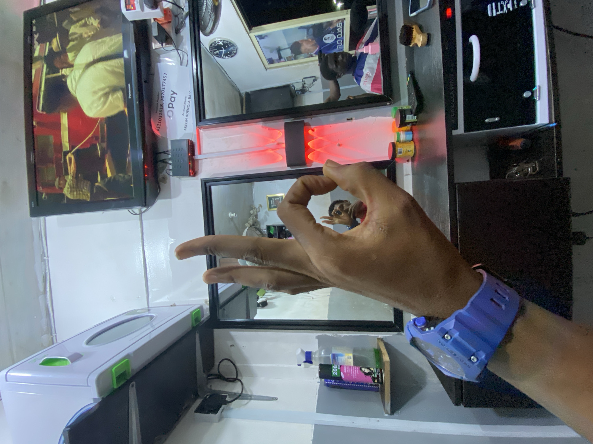 A close-up, vertically oriented image reveals a section of what appears to be a barber shop. A hand with a blue wristwatch is centrally placed in the composition, gesturing with the thumb and forefinger forming a circle. The hand's reflection is visible in a mirror. Behind the hand, various objects are visible, including a black-framed mirror, a TV screen displaying a scene with red accents, and a white sterilizer unit. Bottles and containers sit on shelves below the mirror. Another mirror shows the reflection of a person with a blurred shirt, a clock, and a picture frame. Text on a pay slip is visible, including the account name AKEEM ADENOLA BALO and numbers 6111031618, 7070177457. The OCR text also contains the word Panasonic. There are red lights shining, which illuminate objects. The location appears to be Ado, Nigeria, inferred from the account name on the pay slip.