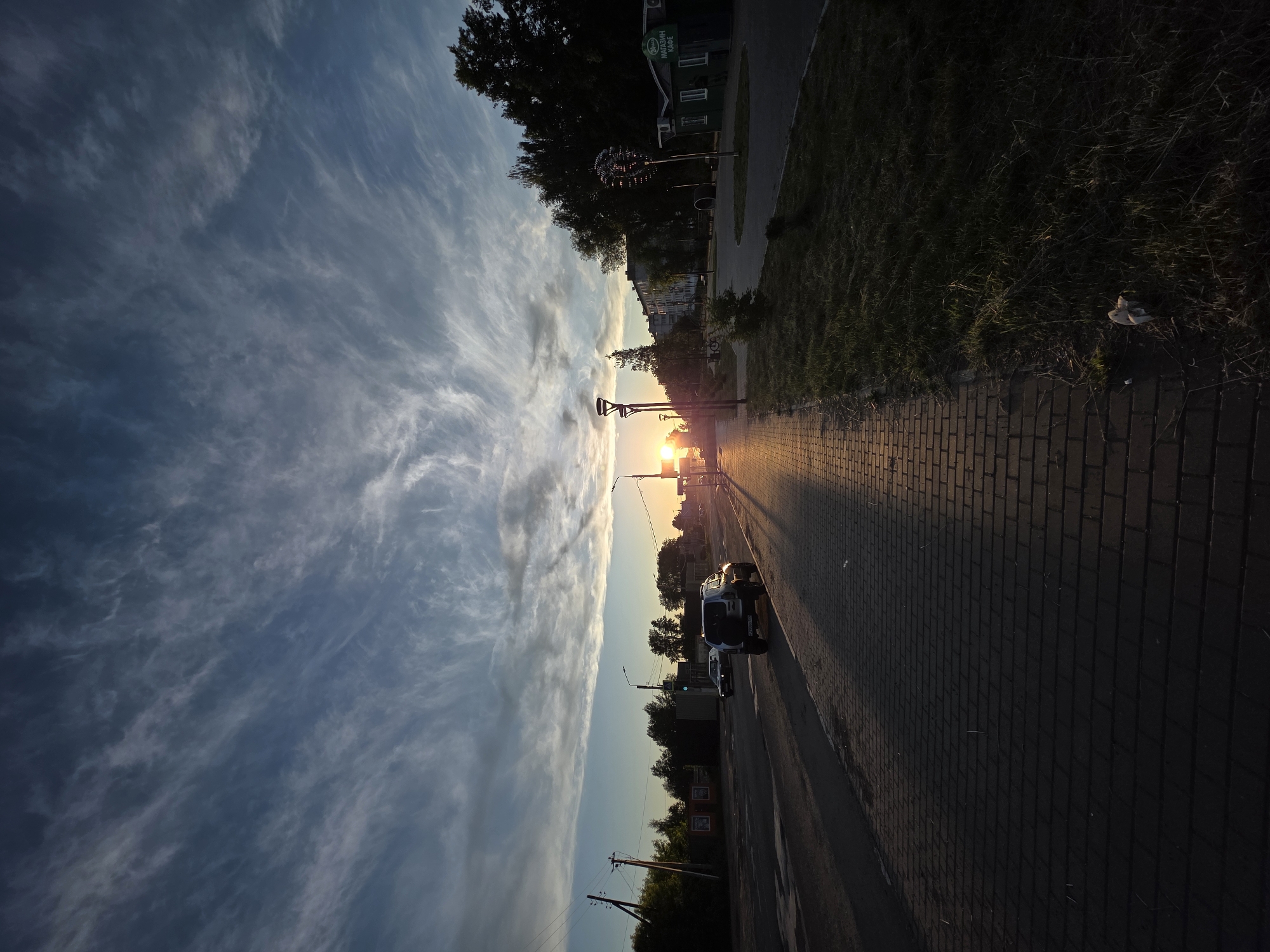 The image depicts a tranquil sunset scene on a street in Chaikovsky, Russia. The sun, low on the horizon, casts a brilliant orange-yellow glow directly into the viewer's perspective, illuminating the street and creating long shadows.
The sky occupies a significant portion of the image, showcasing a striking contrast between the bright, warm hues near the setting sun and the cooler, darker blues and grays higher up and to the left. Wispy, streaky clouds (cirrus) are prominently featured, particularly in the darker sky areas, catching the last light of the day.
At street level, a paved road stretches towards the horizon, with at least two vehicles visible; one appears to be a light-colored SUV or truck parked or moving. A brick-paved sidewalk runs parallel to the road, bordered by a strip of green grass. On the right side, the grass verge also shows some dry patches and a small, discarded white object.
Trees line both sides of the street, with those closer to the sun appearing largely as dark silhouettes. Further back on the left, several buildings are visible, including a dark green building with white windows. This building features a sign with Cyrillic text that reads "МАГАЗИН КАФЕ," translating to "SHOP CAFE." Near this building, a spherical, possibly decorative, metallic object is noticeable. Streetlights and power poles with overhead wires are also present, contributing to the urban or suburban setting.
The overall impression is one of a peaceful evening, with the warm light of the setting sun creating a serene atmosphere.