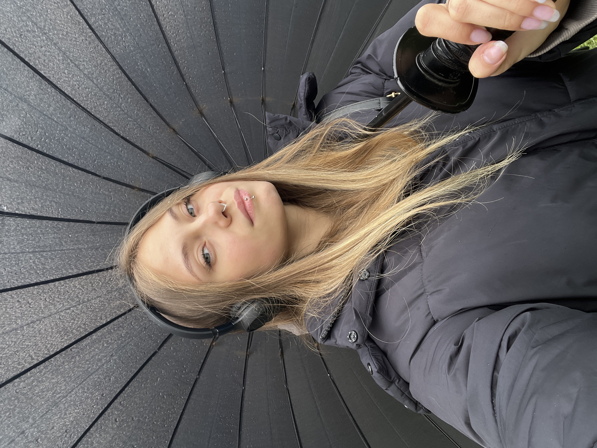 A young, fair-skinned person with light blonde hair is holding a large, open black umbrella. They wear black headphones and a black puffy jacket. The person has piercings in their nose and lip. The scene appears to be outdoors, possibly during or after rain, given the umbrella. There's no information available to determine if the location is Фоки, Russia.