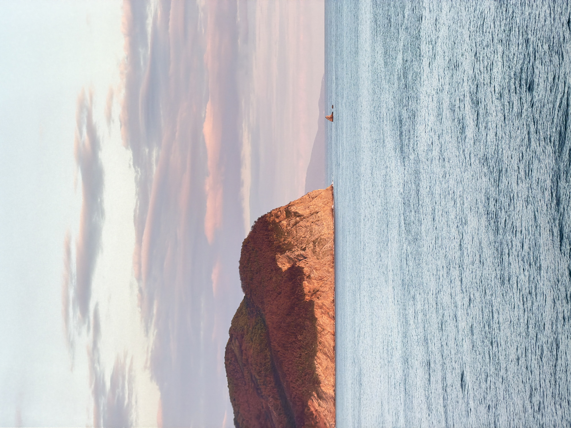 The image presents a rotated landscape scene. On the right side, a body of water stretches out. The water is turquoise in color and the texture appears rough. A rocky coastal promontory occupies the left. The rock is predominantly brown and slopes down towards the sea. The sky above is colored with hues of light pink, grey, and blue with streaks of cloud formations. In the horizon, a small object resembling a sailing boat is seen in the sea. This depicts the scenery around Petrovac na Moru, Montenegro.