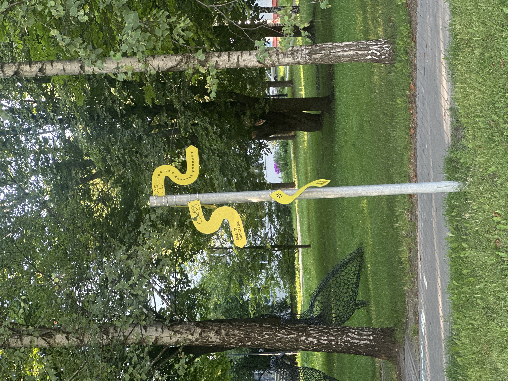 The image shows a metal pole with several yellow signs in a park-like setting. The signs are shaped like numbers and contain Russian text and symbols. One sign shows the number "2" with rings, and text that translates to "About family". Another sign shows an arrow pointing left with text that translates to "Together forever". A third sign shows "18+" with a wavy line. The pole is situated on a paved path, with a grassy area and trees surrounding it. In the background, there are buildings and some decorative wire sculptures of birds. The setting appears to be a public park in Minsk, Belarus, likely a recreational area designed for families.