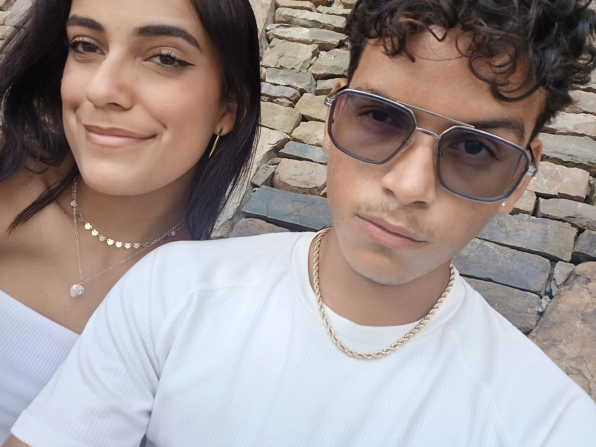 Two people, likely a young adult female and male, pose for a selfie. The female is on the left, wearing a white top and two necklaces. Her hair is dark, and she has winged eyeliner. The male is on the right, wearing a white t-shirt, a gold chain, and aviator-style sunglasses. He has curly hair. The background is a stone wall. The location is identified as Juan Griego, Venezuela.