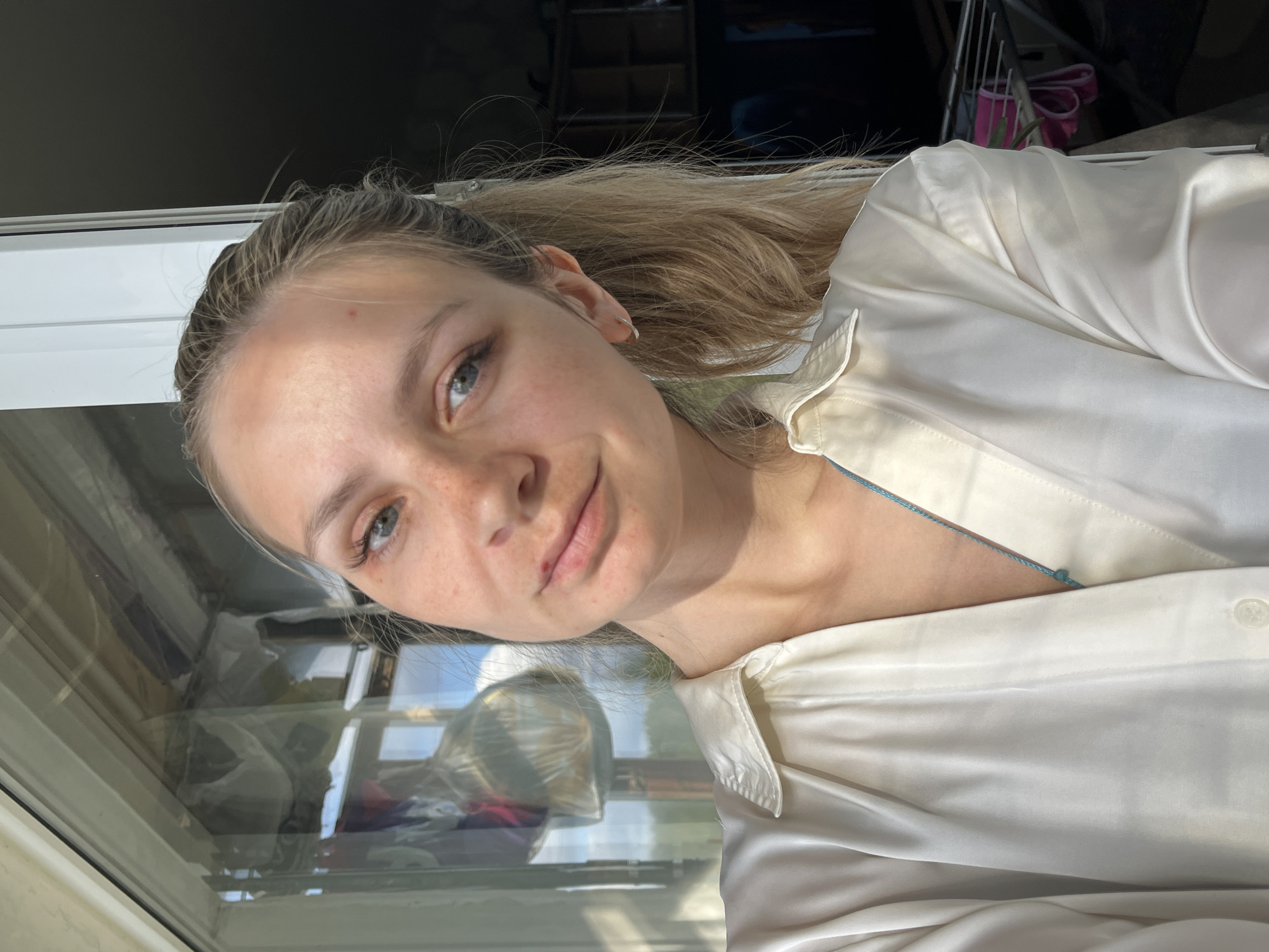 A selfie image shows a young woman with fair skin, blonde hair pulled back in a low ponytail, and blue eyes. She is smiling slightly and looking directly at the camera. She is wearing a white, possibly silk, button-down shirt. The background is somewhat blurred, but it appears she is indoors, possibly near a window. Reflections in the window suggest an outdoor environment. There is no specific information available to indicate that the photo was taken in Chaikovsky, Russia.