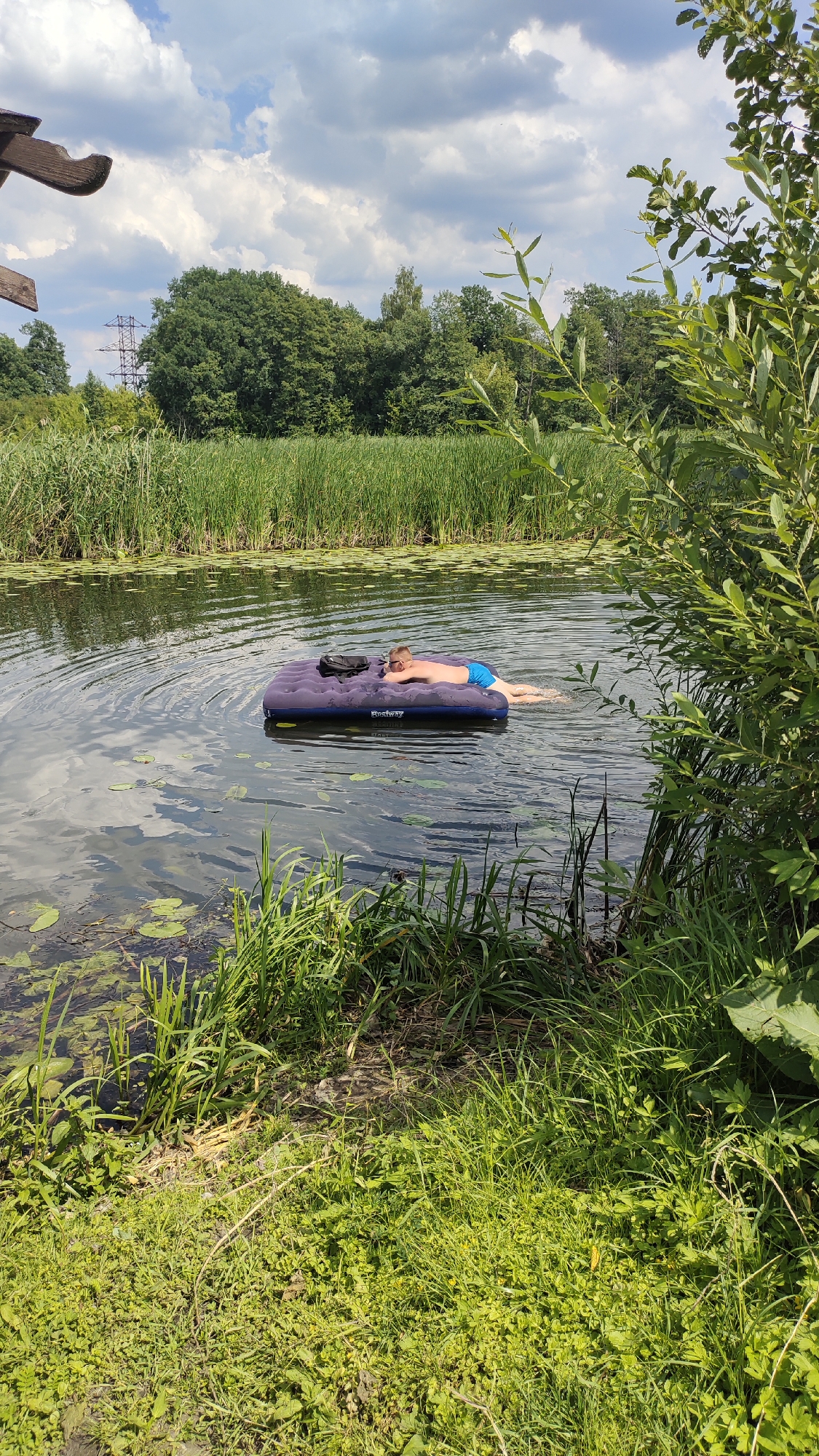 A young male wearing blue swim trunks is lying on a blue inflatable mattress in a body of water. He is face down, with his head turned to the left, and his arms extended. The mattress has the brand name "Bestway" visible on it. The water is calm, with ripples emanating from the mattress. Lily pads are visible on the surface of the water, especially in the foreground and to the left of the mattress. The background consists of tall green reeds lining the bank of the water, followed by a dense forest of green trees. Above the trees, a cloudy sky is visible. To the left side of the image, a wooden structure with a curved edge is partially visible. The scene appears to be a natural outdoor setting, possibly a river or lake, in Ramon, Russia.