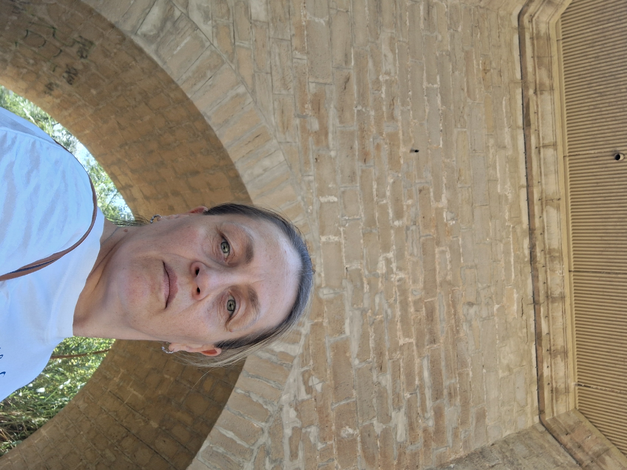 A medium shot captures a middle-aged Caucasian woman from the chest up, looking directly at the camera. She has light skin, blonde hair pulled back, and is wearing a white t-shirt and hoop earrings. The background consists of a stone archway and a brick wall, suggesting an outdoor location. The lighting is bright, indicating daytime. The scene is set in Alcañiz, Spain.