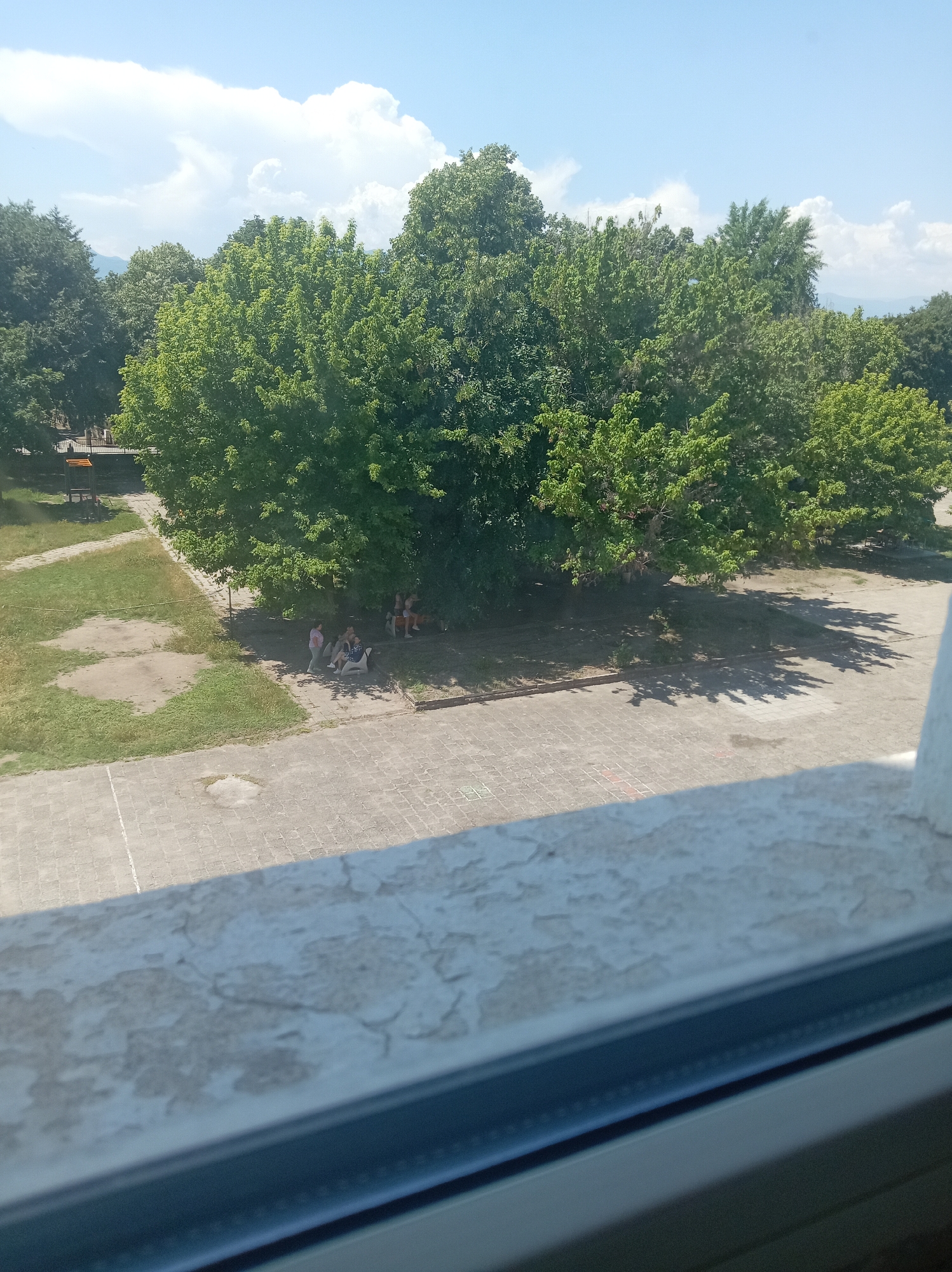The image shows an outdoor scene on a sunny day in Stamboliyski, Bulgaria. In the foreground, a concrete ledge with peeling paint is visible, suggesting it is part of a window sill or balcony railing. Beyond this, a paved area with a grid pattern, likely a courtyard or sidewalk, is seen. To the left of the paved area is a patch of green grass with some bare earth spots. Further into the scene, a group of people, appearing to be children and adults, are gathered under the shade of large, lush green trees. One person is wearing a pink shirt and dark pants, and others are seated on benches or the ground. The trees form a dense canopy, casting shadows on the ground. In the background, more trees are visible, and beyond them, hazy hills or mountains can be seen against a bright blue sky with scattered white clouds. There is also some playground equipment visible in the far left background. The overall impression is of a recreational area or park on a warm day.